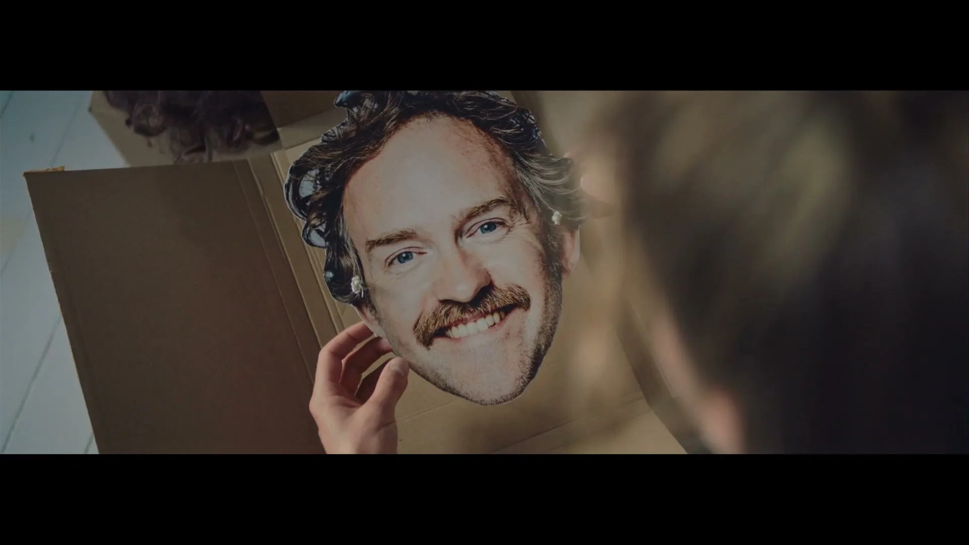 A person's hands hold up a cardboard cutout of a smiling man's face with a mustache, seen from an over-the-shoulder perspective.