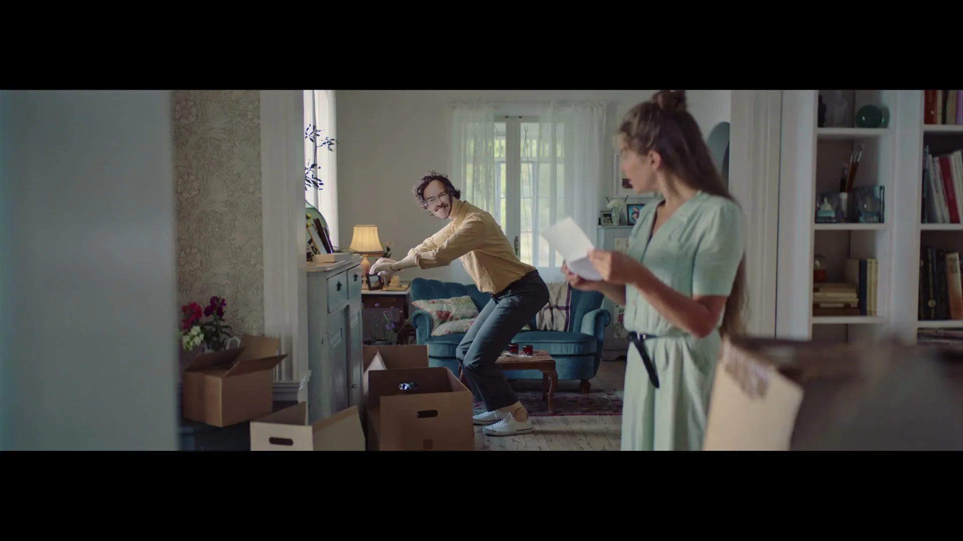 A man in a yellow shirt and glasses smiles at a woman in a green dress who is reading a letter in a living room with moving boxes.