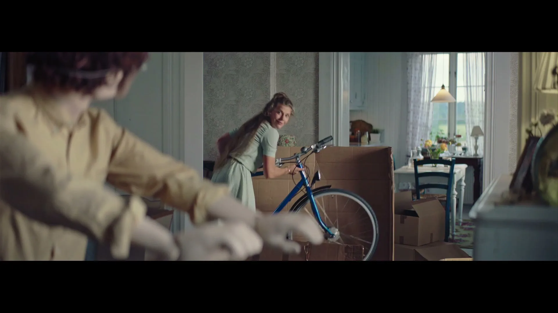 A young woman in a light green dress smiles at a doll in the foreground while assembling a blue bicycle in a living room filled with moving boxes.