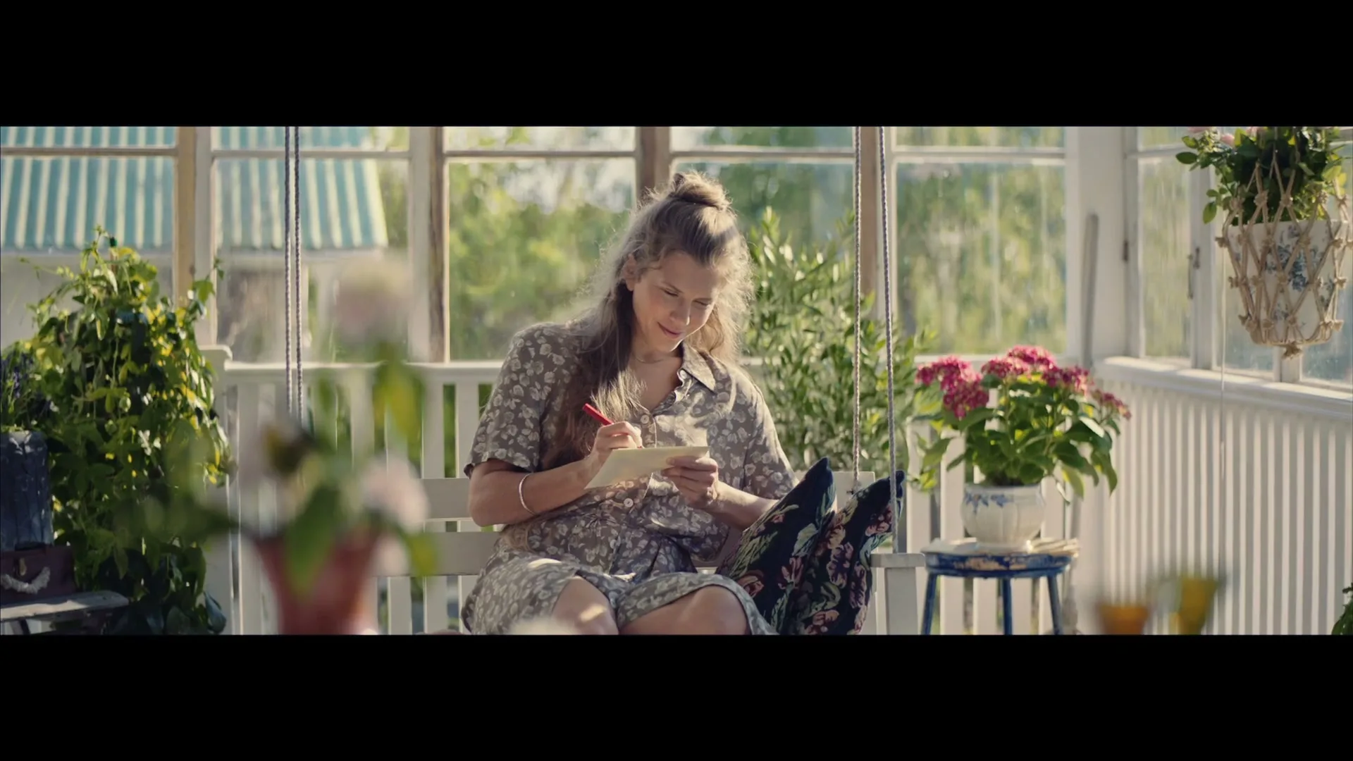 A young woman with light brown hair in a floral dress sits on a porch swing, writing a letter in a sunlit conservatory.