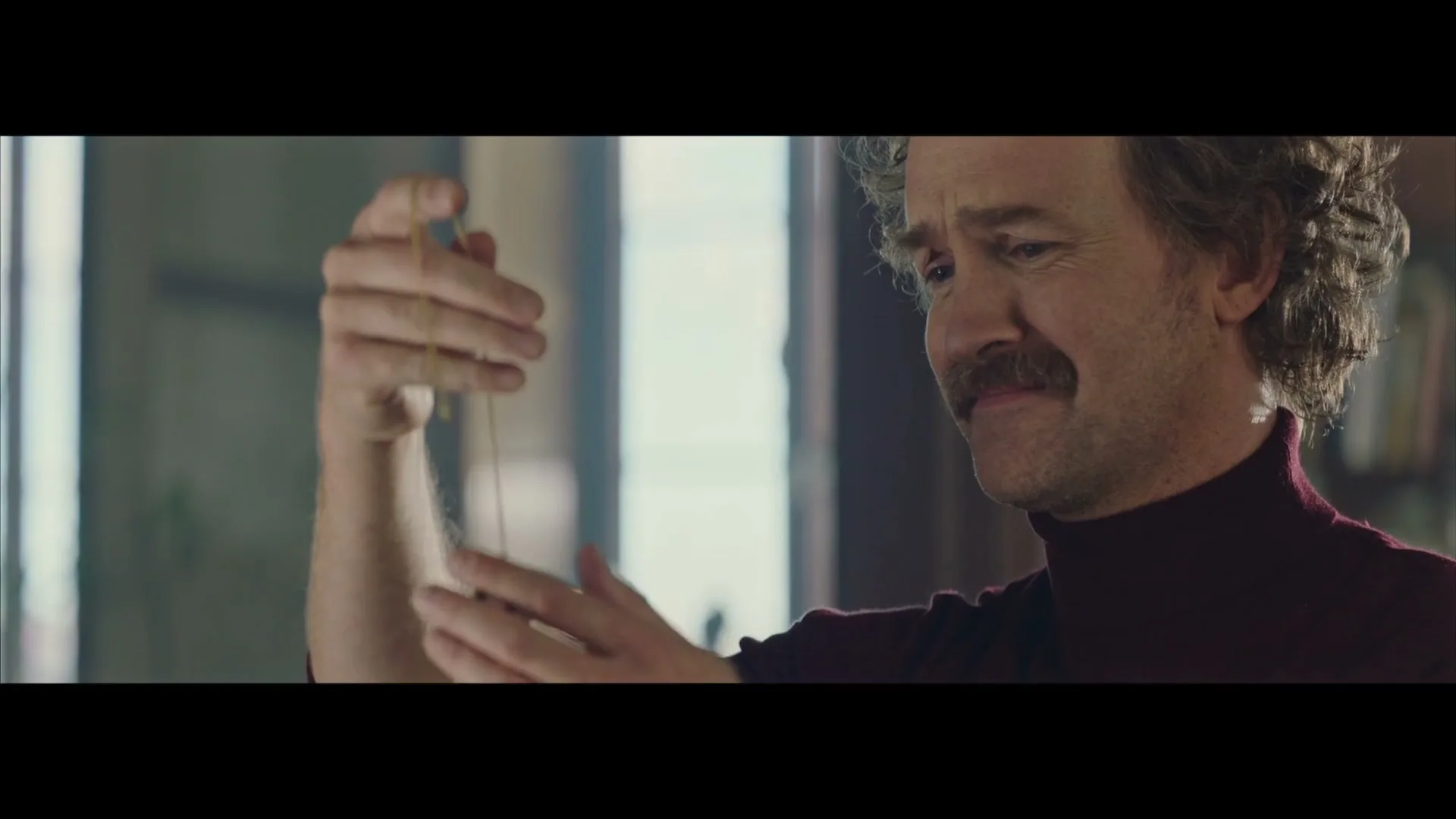 A man with a mustache and curly hair, wearing a turtleneck, holds a locket on a chain, looking at it with a sad expression indoors.