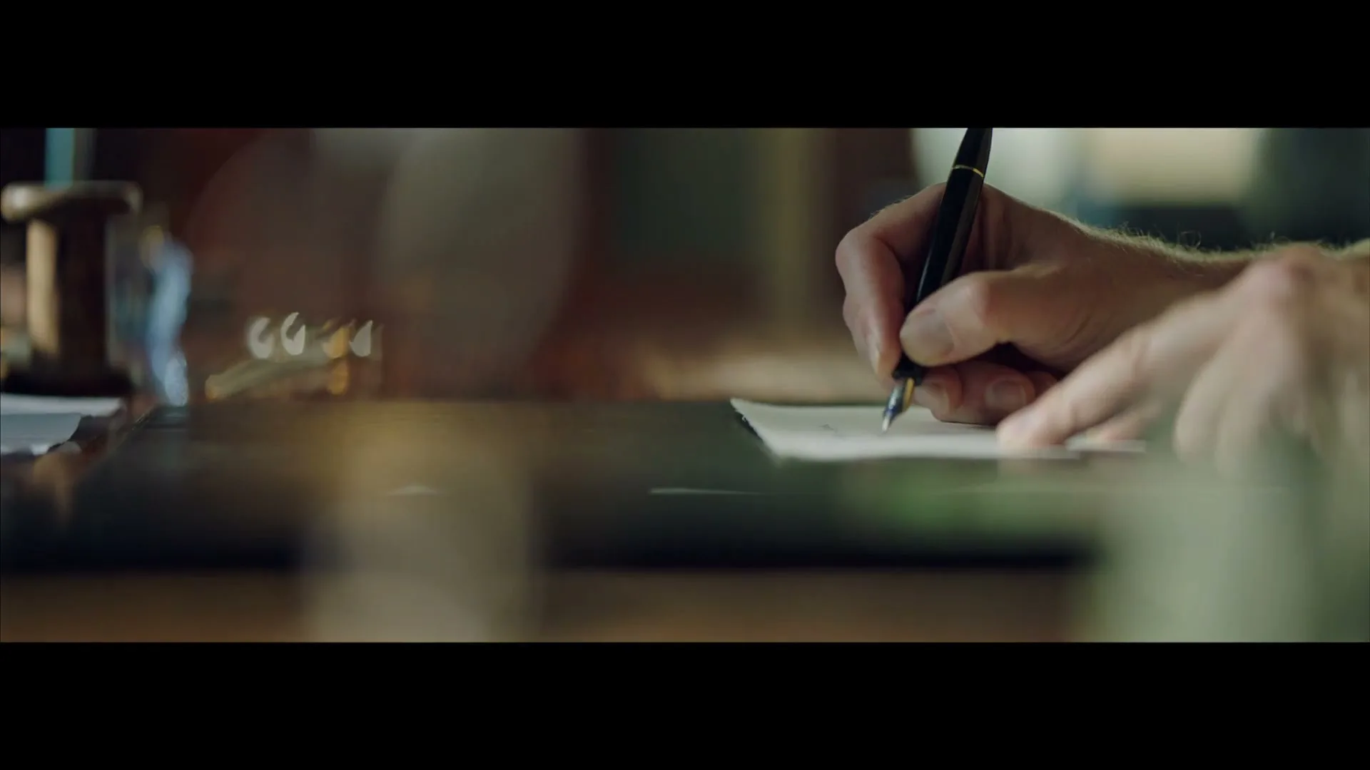 A man's hands are seen in a medium closeup, writing a letter with a fountain pen on a dark wooden desk in an interior setting.