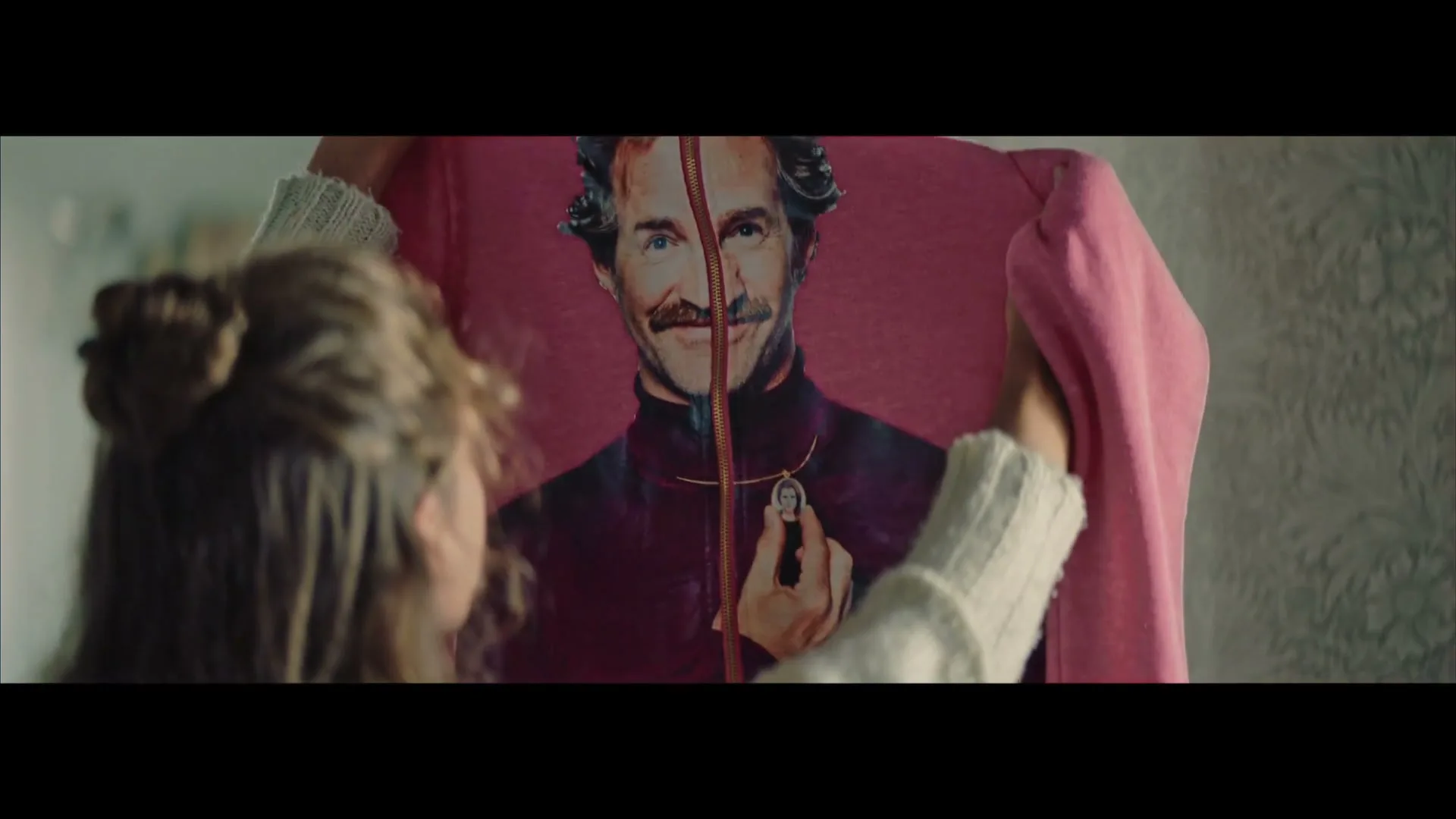 A young woman with her hair in a bun holds up a pink t-shirt with a man's face printed on it, in a softly lit room.