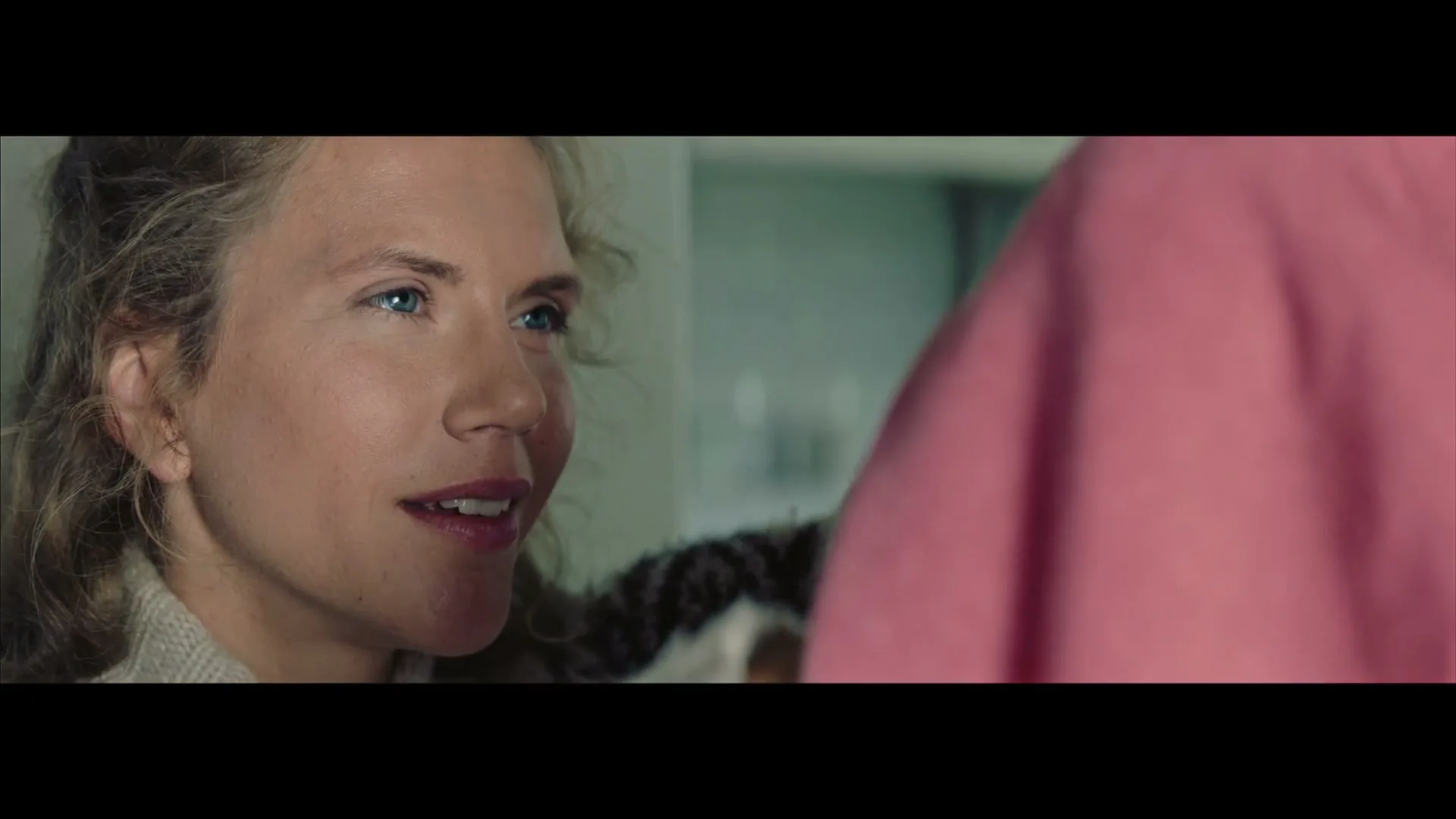 A woman with blonde curly hair and blue eyes looks at someone off-screen, smiling softly in a domestic interior.