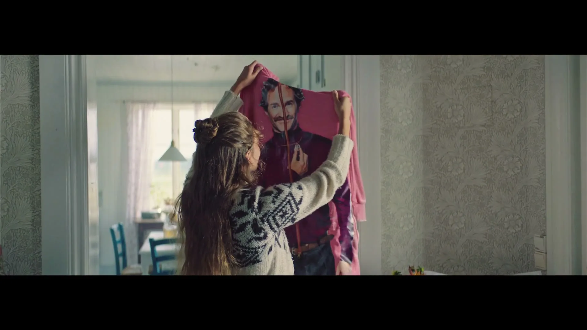 A young woman with long brown hair in a patterned sweater holds up a pink onesie with a man's face printed on it, in a bright, cozy room.