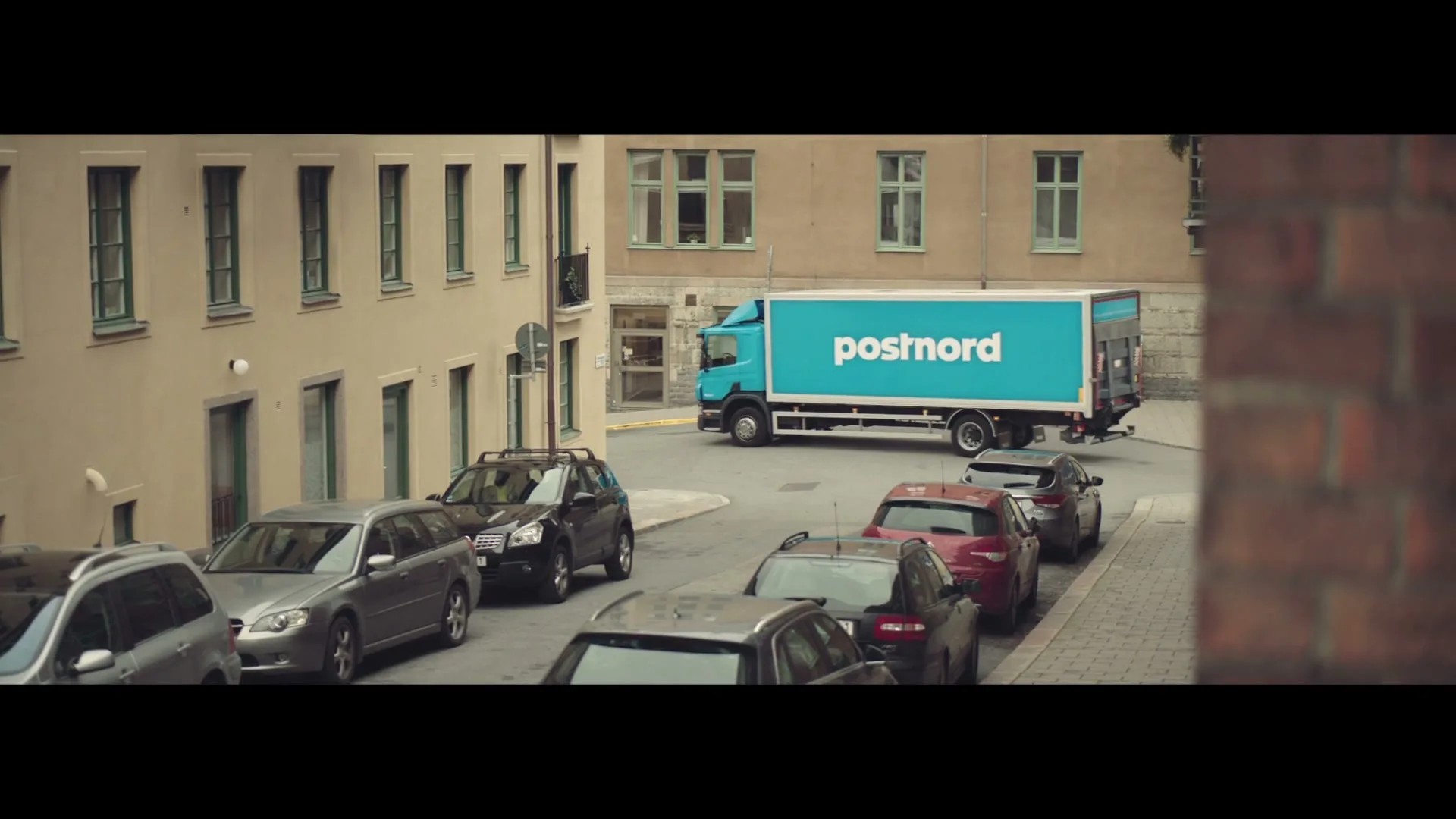 A blue Postnord truck drives through a city street lined with parked cars and buildings, with a brick wall in the foreground.