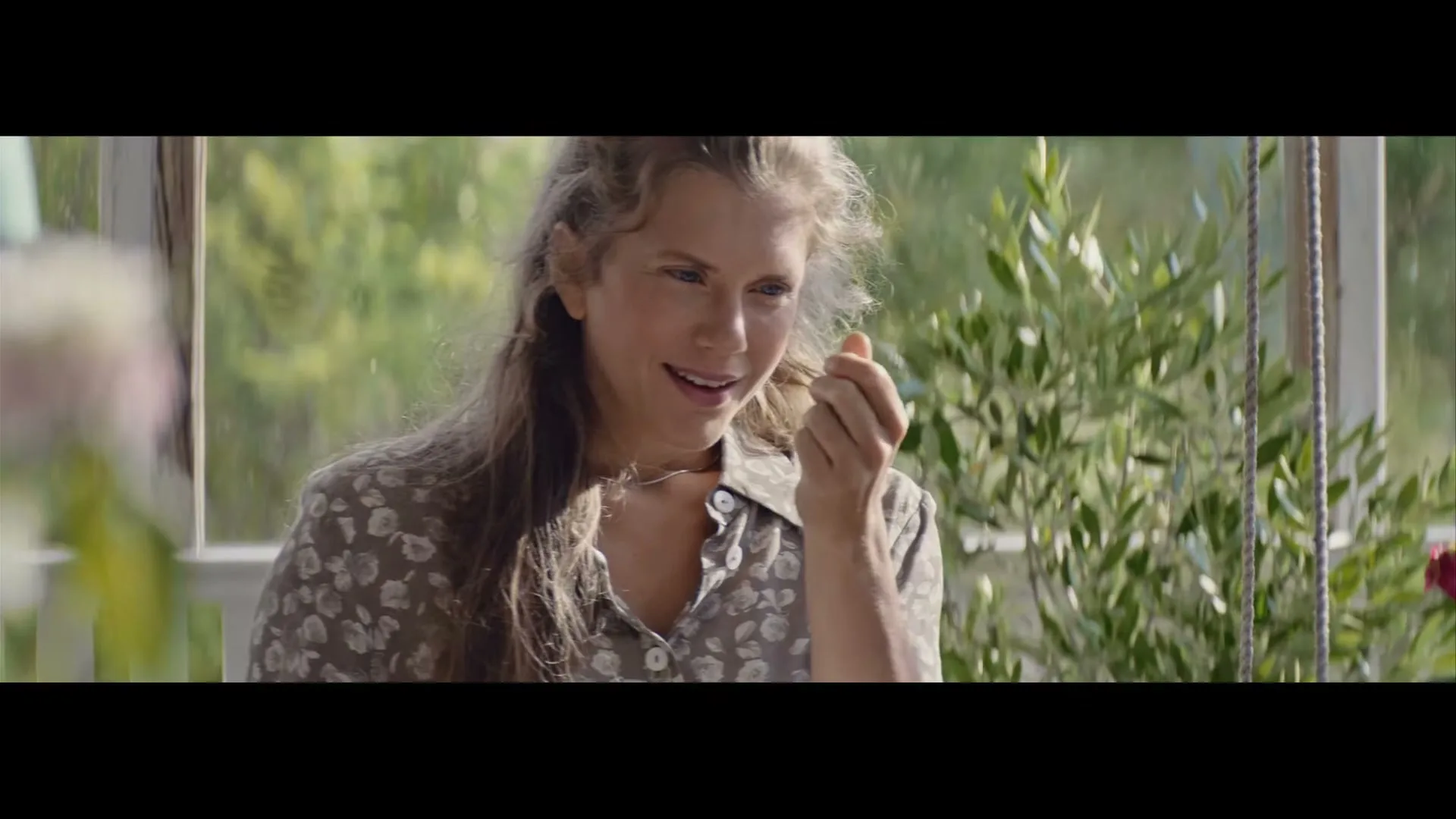 A young woman with long, wavy hair, wearing a floral shirt, smiles while looking down and holding something small in her hand on a sunny porch.