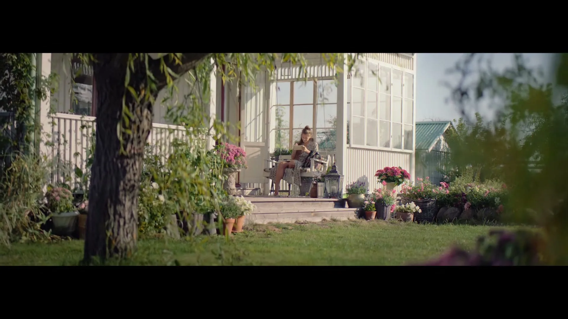 A woman sits on the porch of a white house, surrounded by lush green garden and flowers, reading a letter.