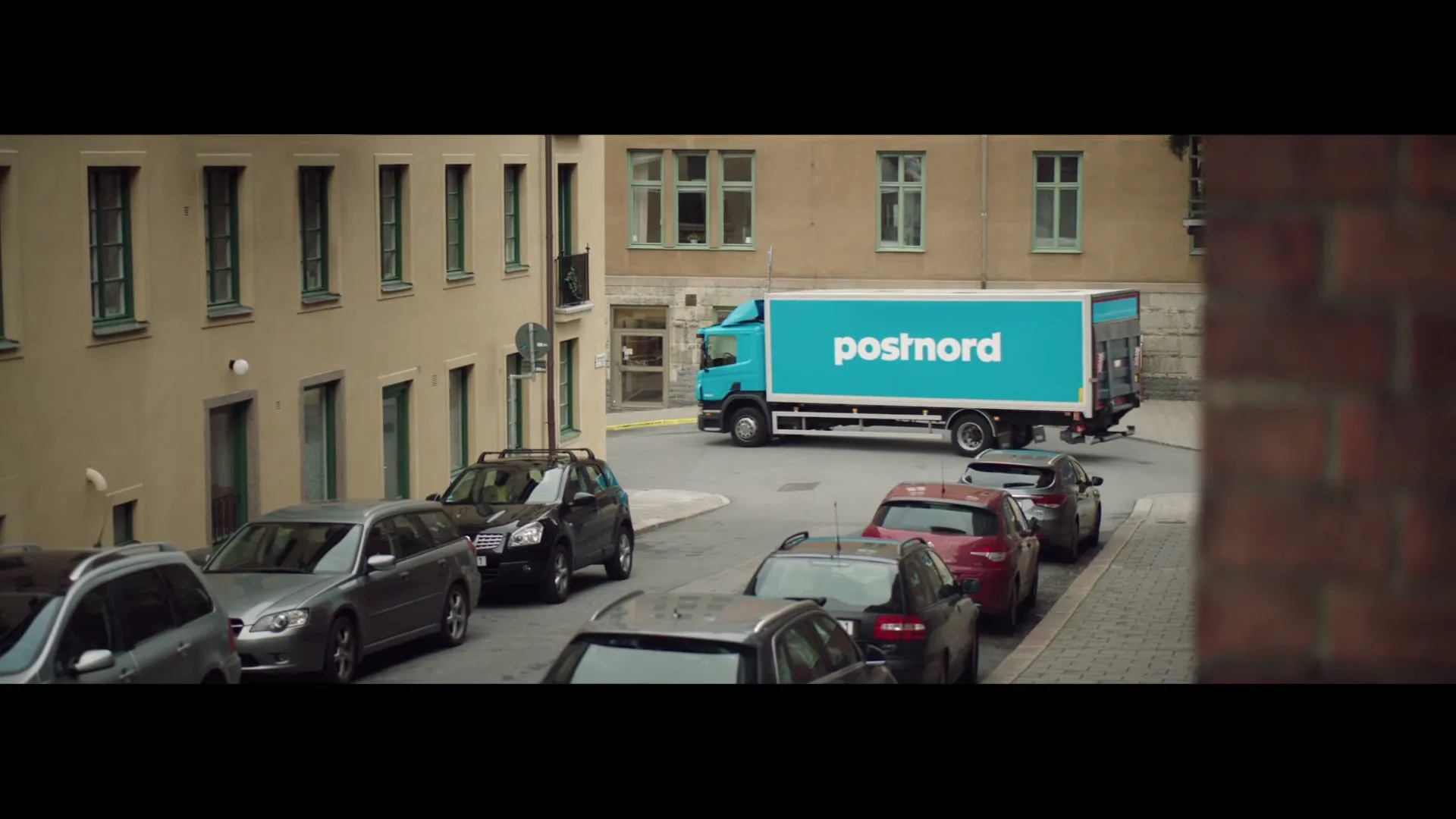 A blue Postnord truck drives down a narrow street lined with parked cars and apartment buildings on a cloudy day.