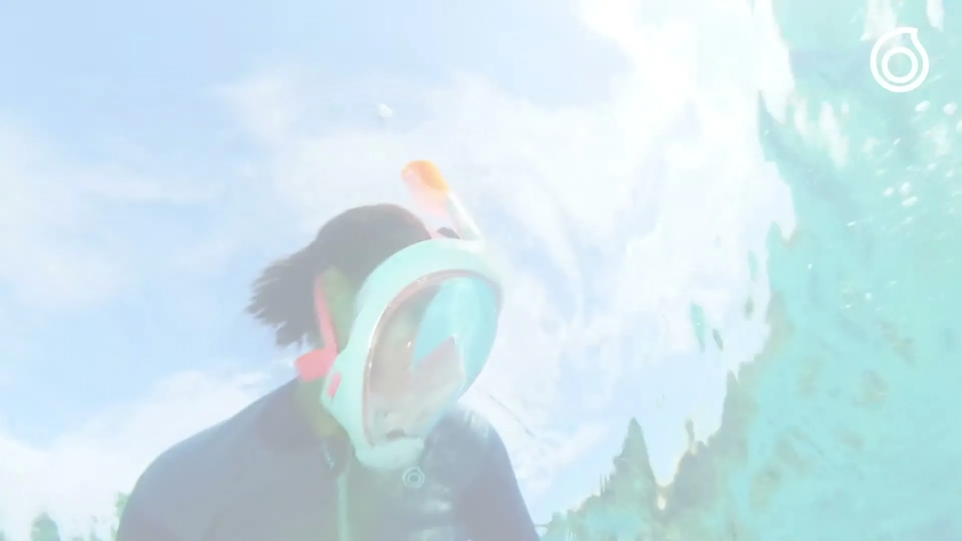 A person wearing a full-face snorkeling mask is seen from below the water, looking up at the bright sky and rippling surface.