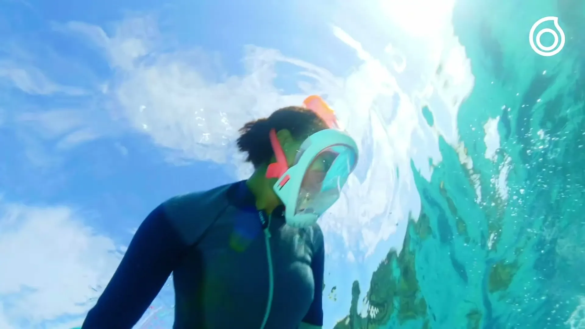 A person wearing a full-face snorkel mask swims underwater, looking up at the bright surface of the ocean.