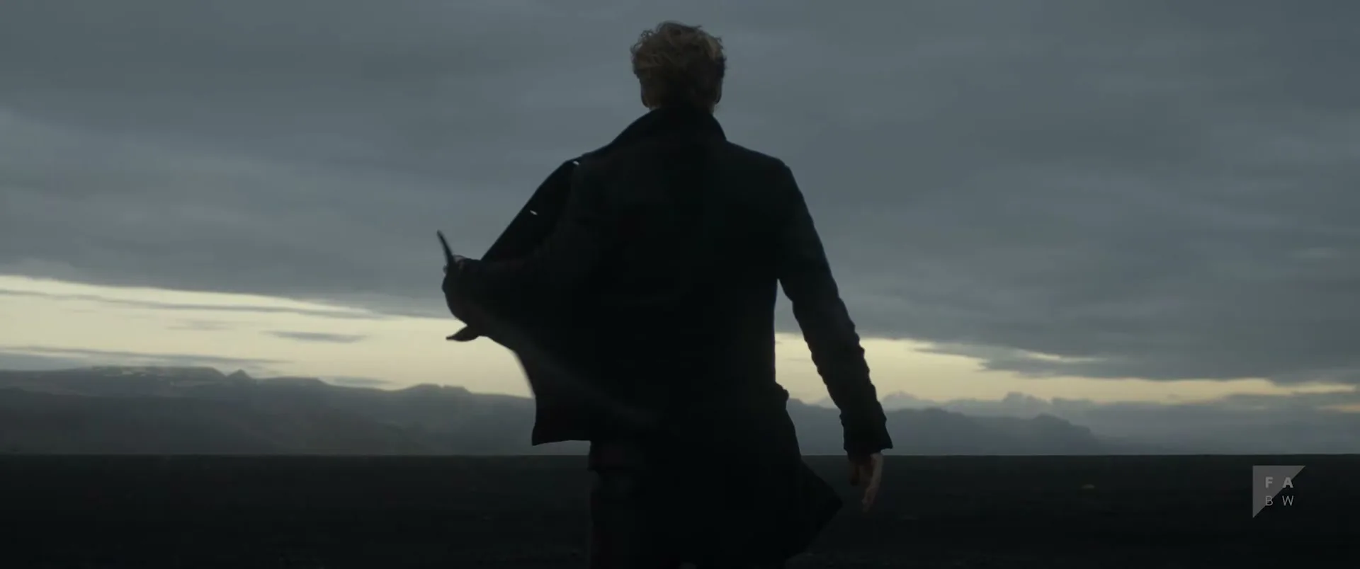 A man with blonde hair and a dark coat stands with his back to the camera, looking out at a vast, dark landscape under a cloudy sky.