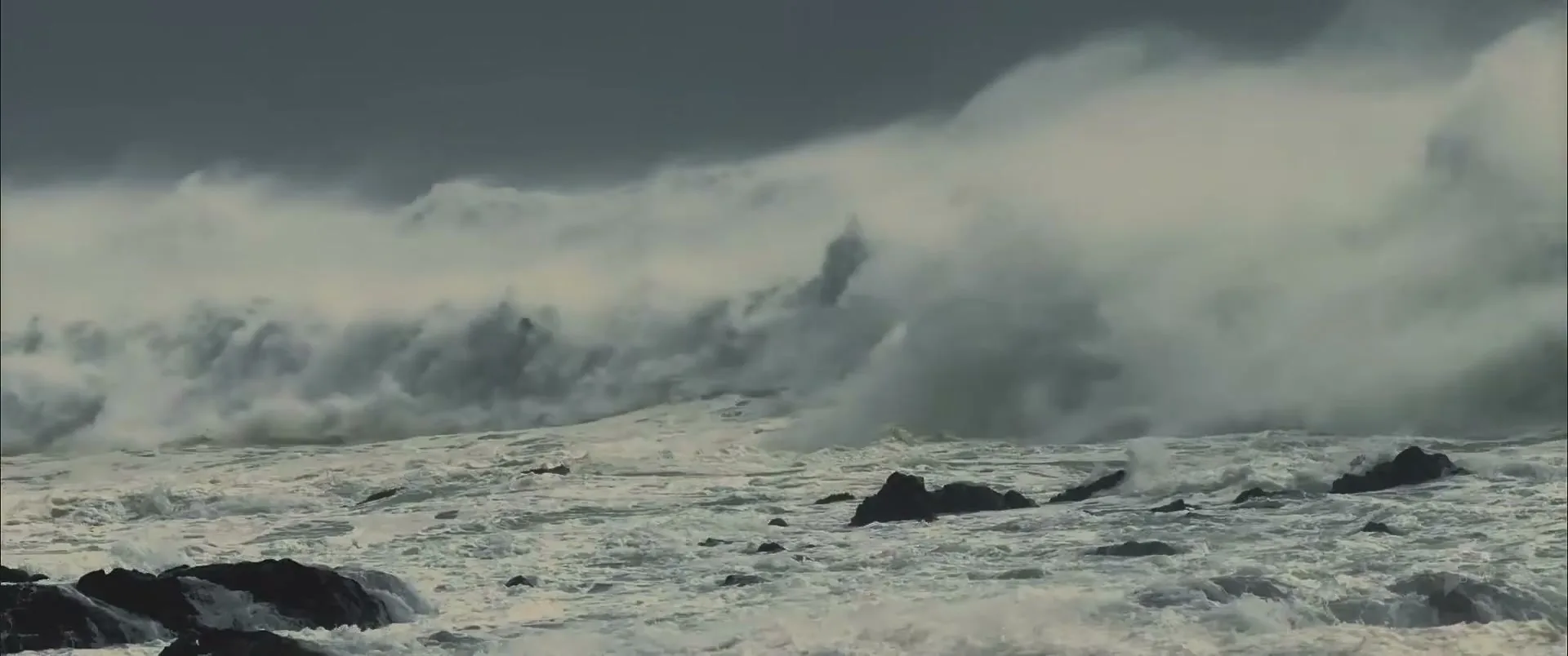 A wide shot captures massive, powerful waves crashing against dark rocks on a stormy day, creating a dramatic and intense scene.