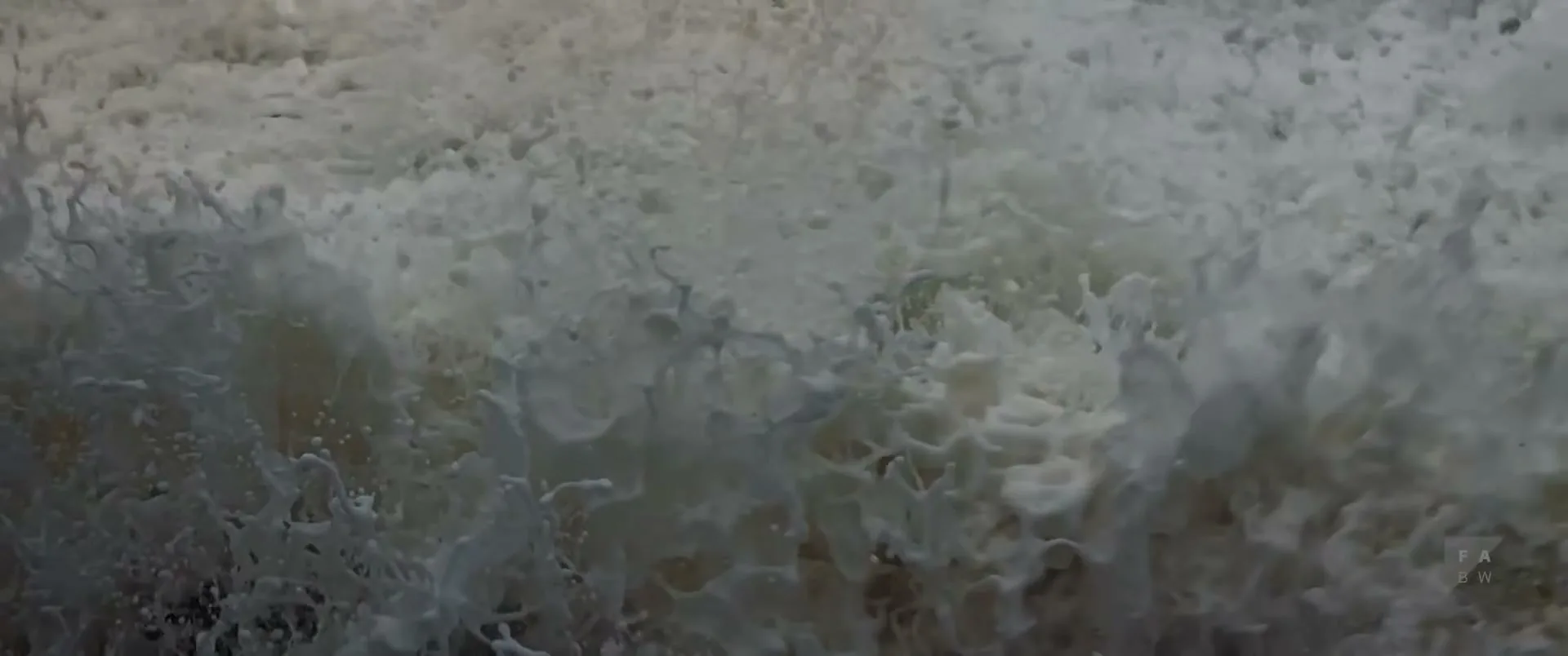 A close-up shot shows the turbulent, foamy surface of a powerful wave crashing, with white spray and bubbles filling the frame.