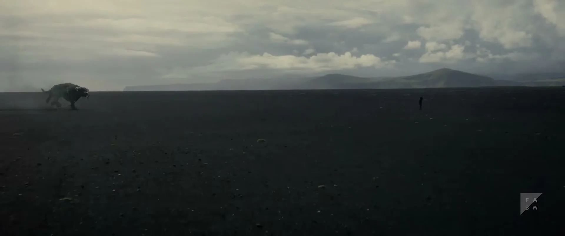 A lone figure stands on a vast black sand beach as a large, dark, wolf-like creature runs towards them under a cloudy sky.