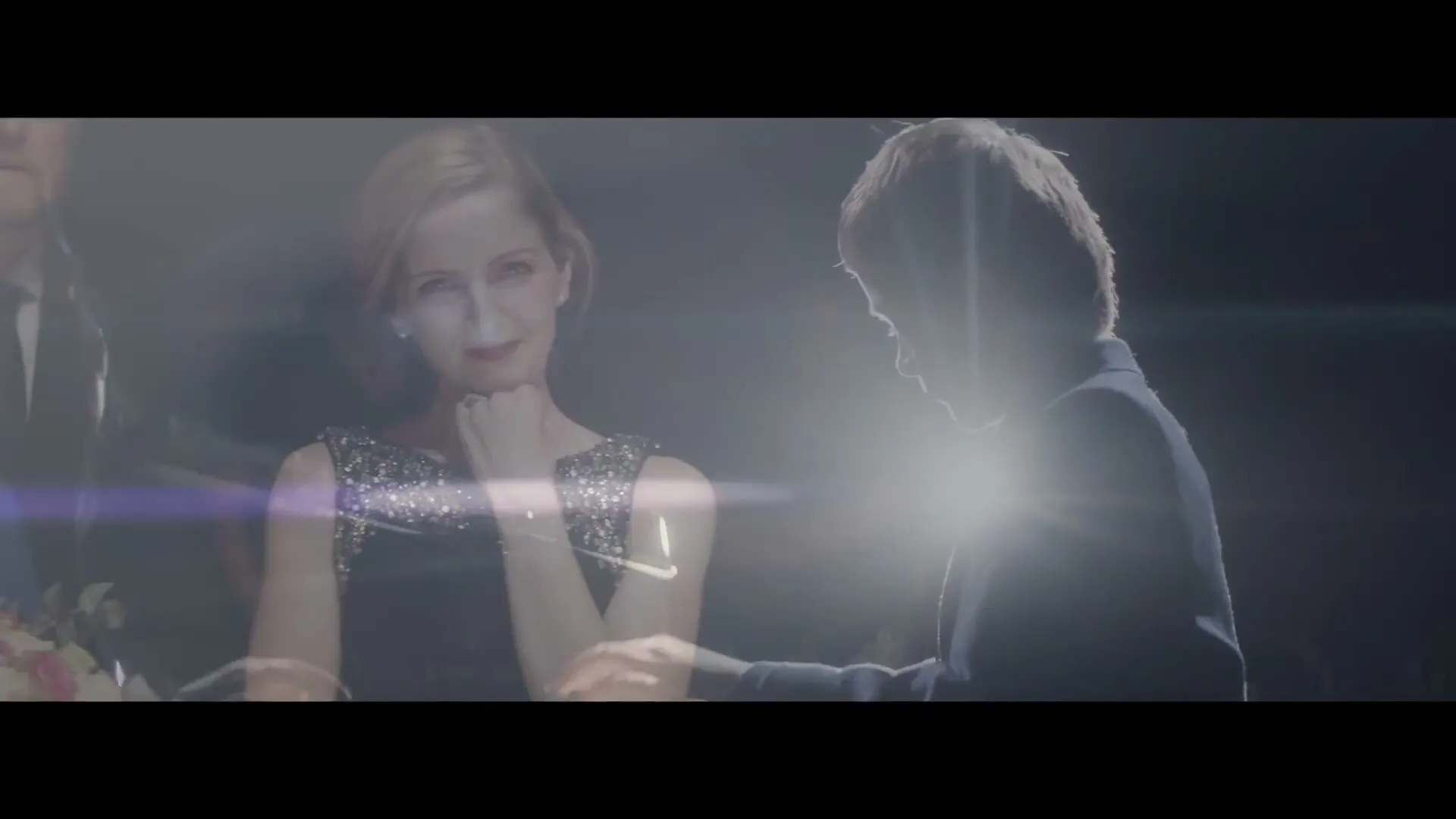 A woman in a sparkly dress smiles while a man in a suit is silhouetted by a bright light, creating a lens flare.