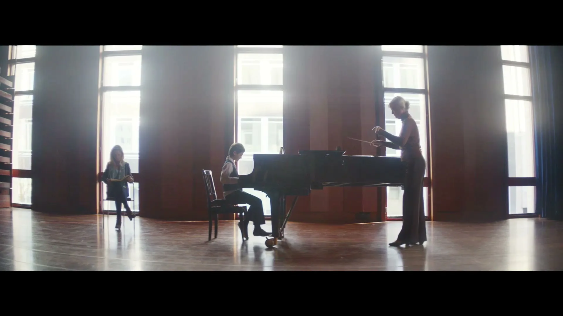 A young boy plays a grand piano while a woman conducts him, and another woman watches from a distance in a bright, spacious room with large windows.