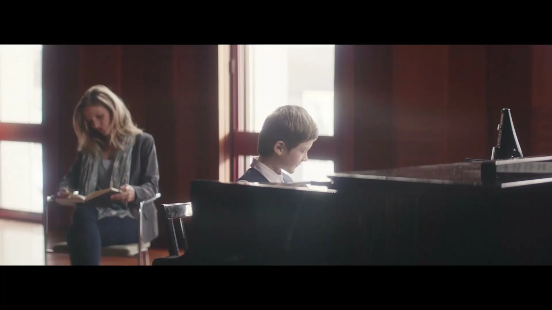 A young boy with light brown hair plays the piano while a woman with blonde hair reads a book in a sunlit room with wood-paneled walls.