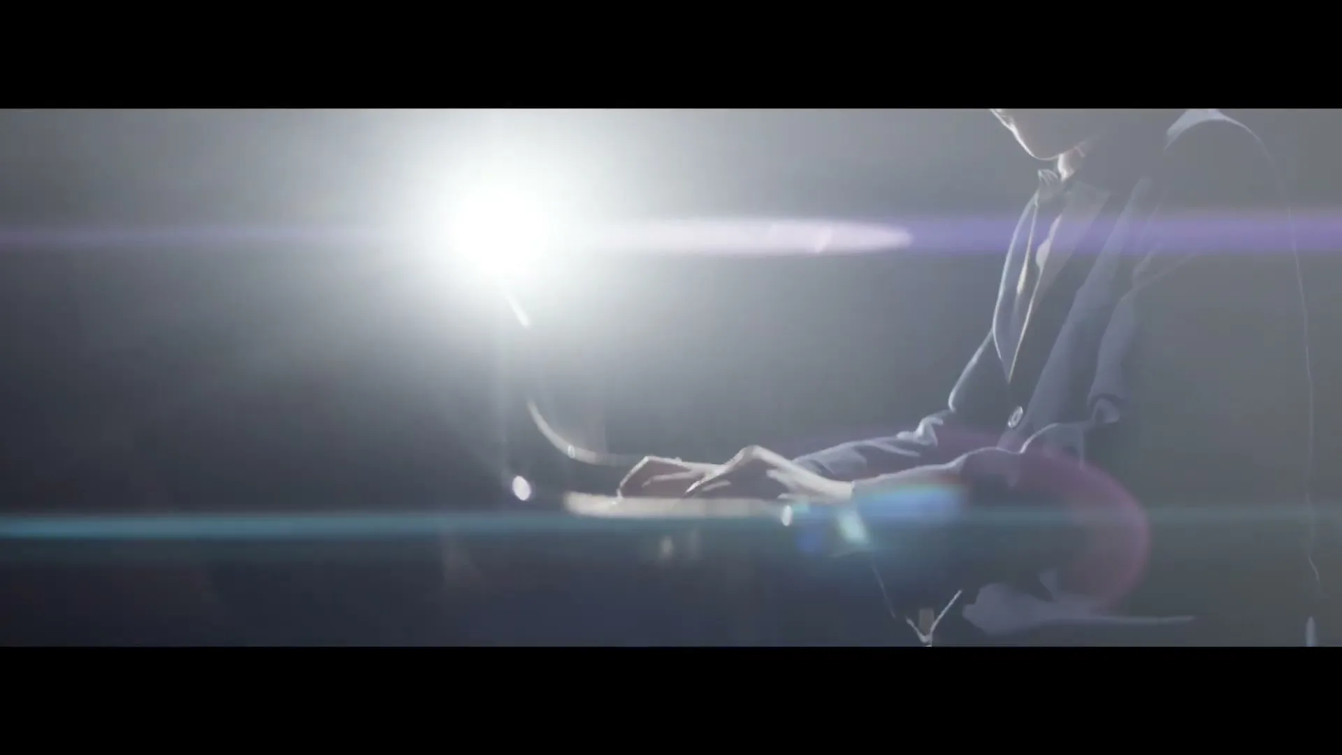A young man in a suit plays a piano, illuminated by a bright spotlight and lens flare in a dark, indeterminate interior.