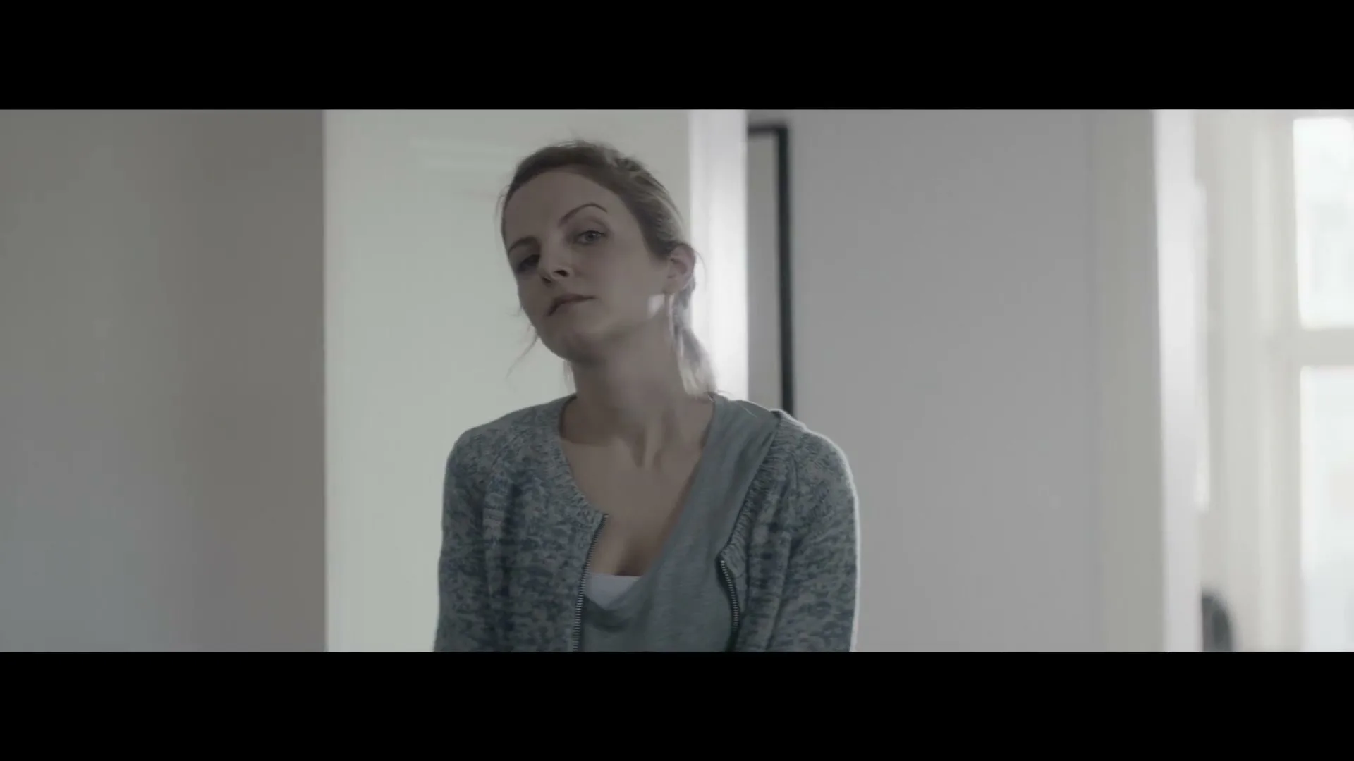 A woman with light brown hair in a grey cardigan and t-shirt looks around a bright, minimalist room.