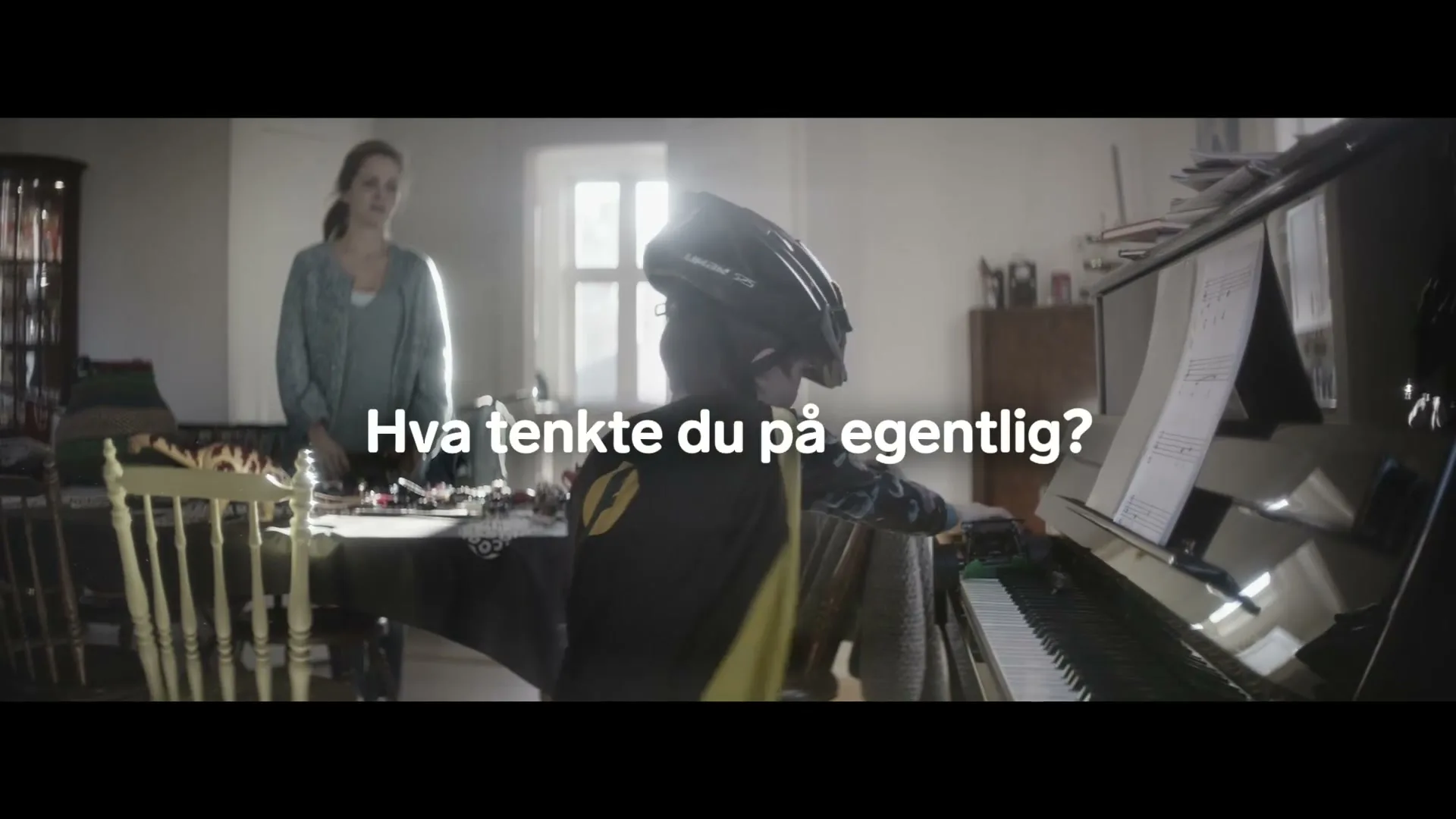 A young boy wearing a bike helmet and cape plays the piano loudly, while a woman stands behind him, looking on in a sunlit room.