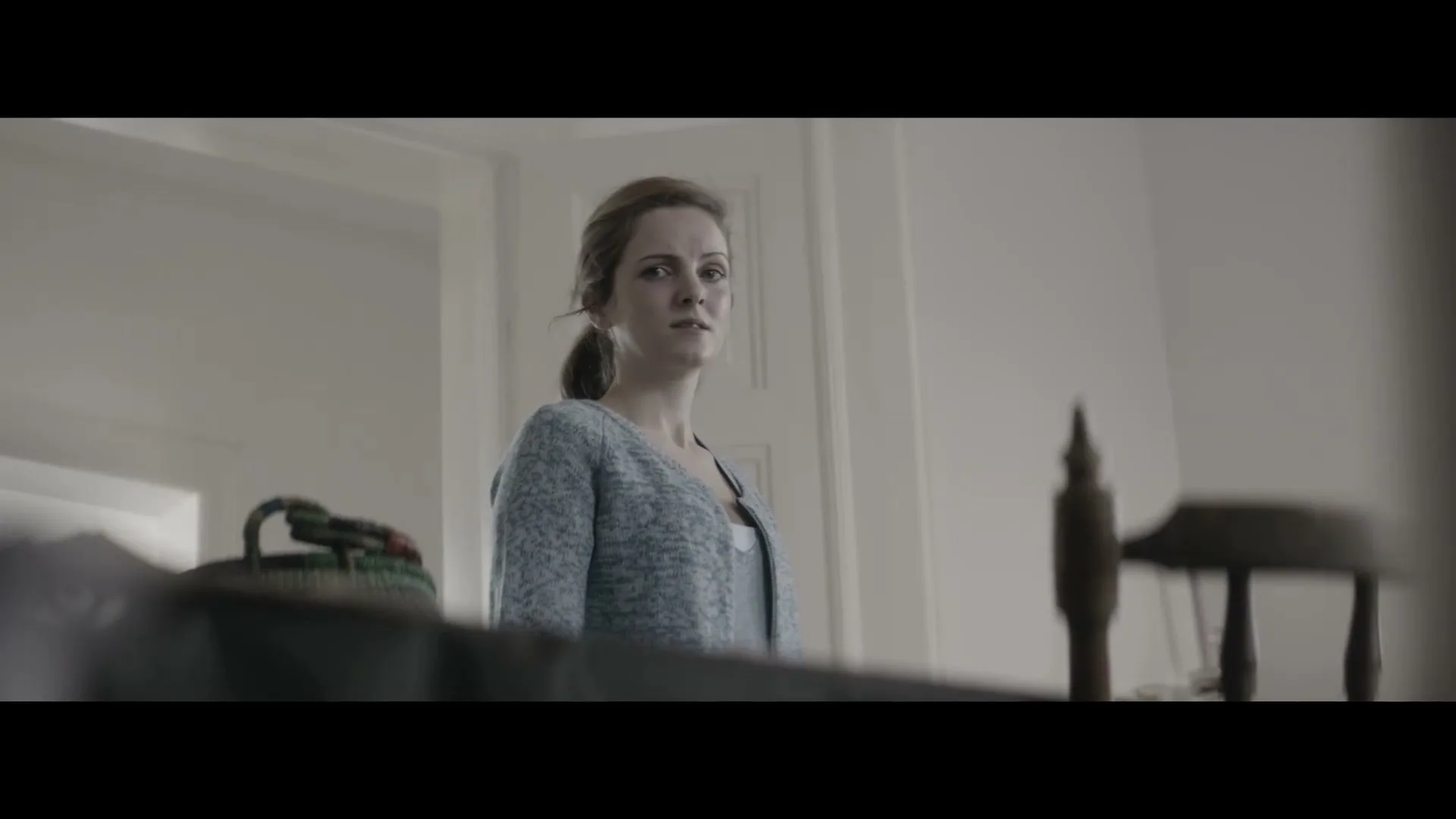 A young woman with brown hair in a ponytail, wearing a light blue cardigan, looks concerned in a brightly lit room.