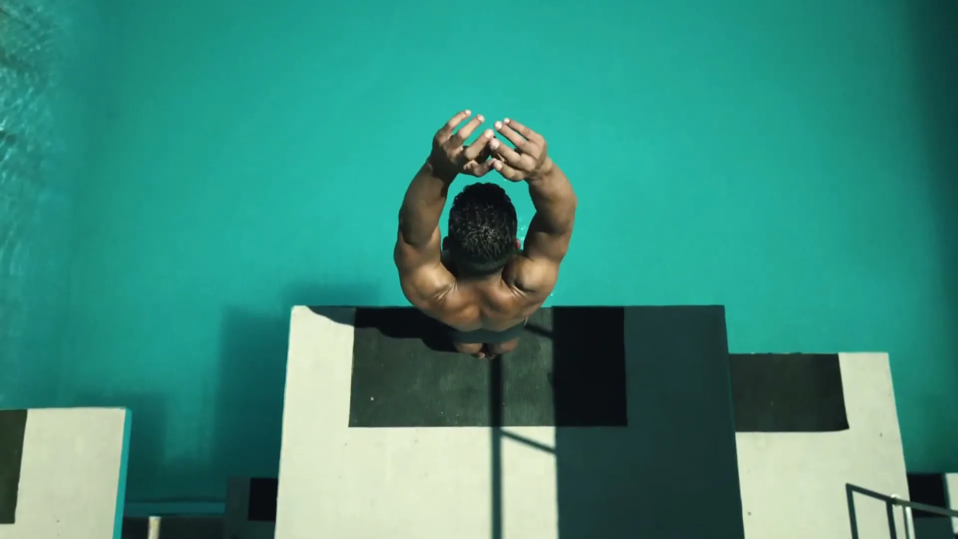 A male diver stands on a diving board, stretching his arms above his head, preparing to dive into a turquoise pool.