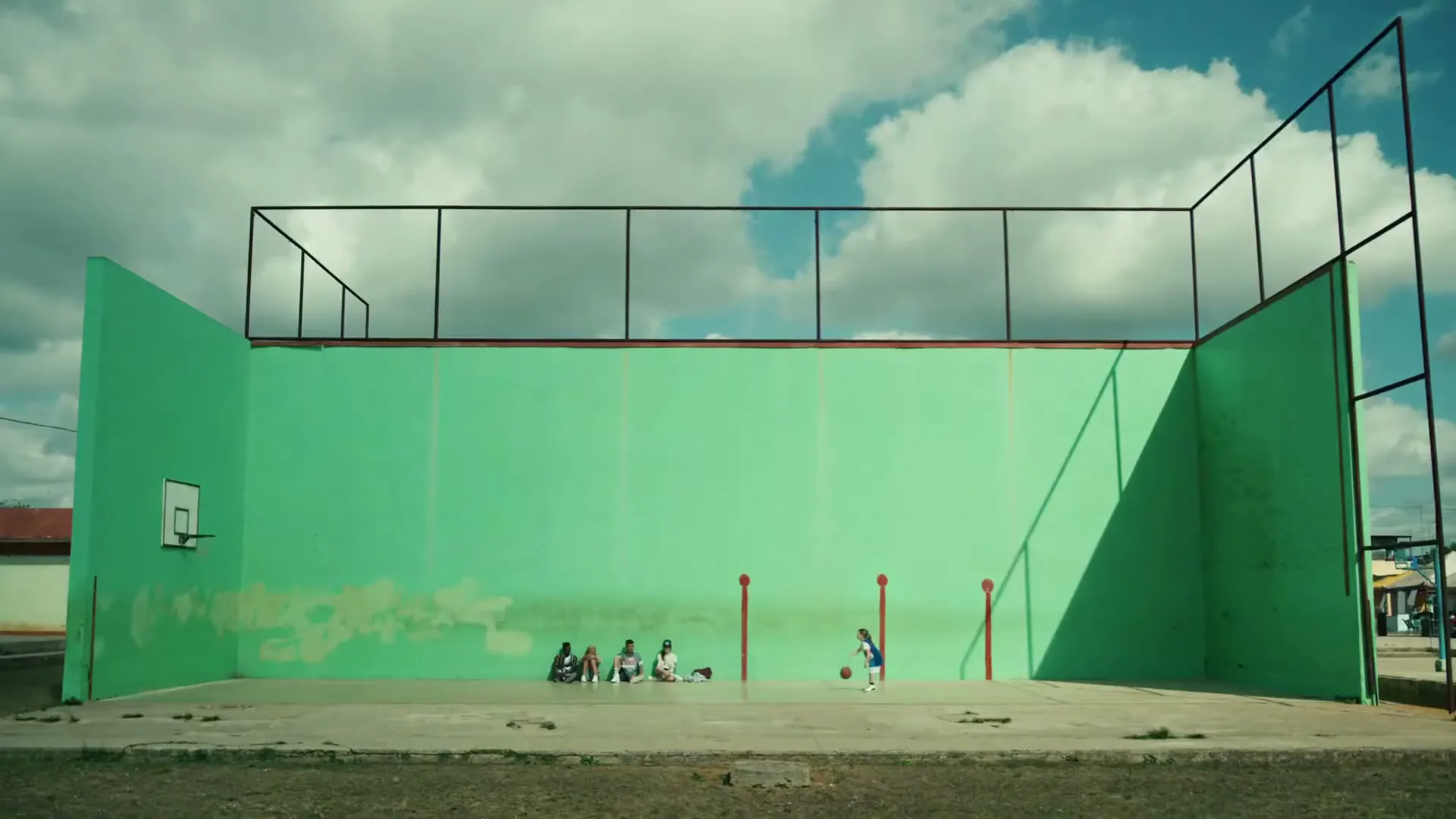 A wide shot shows a young boy dribbling a basketball on a green court while four other children sit on the ground watching him under a cloudy sky.