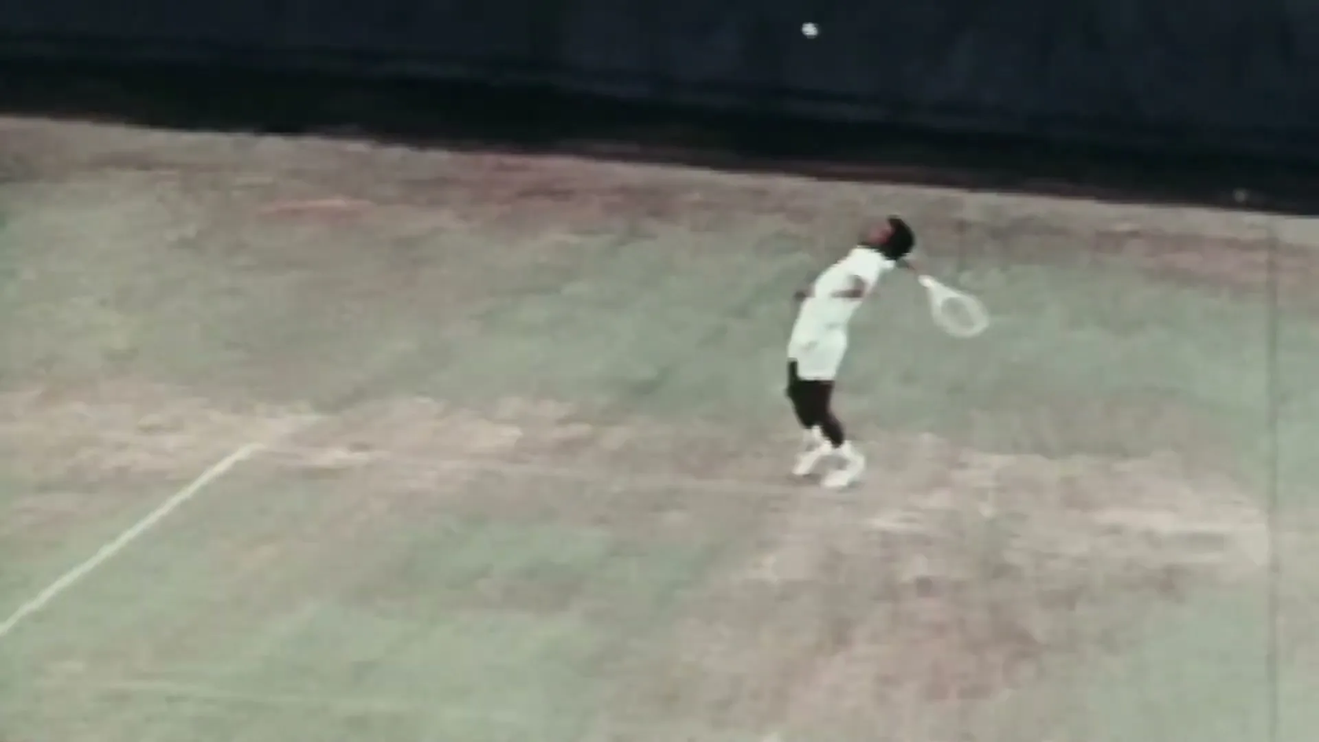 A male tennis player in white sportswear serves a tennis ball on a green court, captured from archival footage.