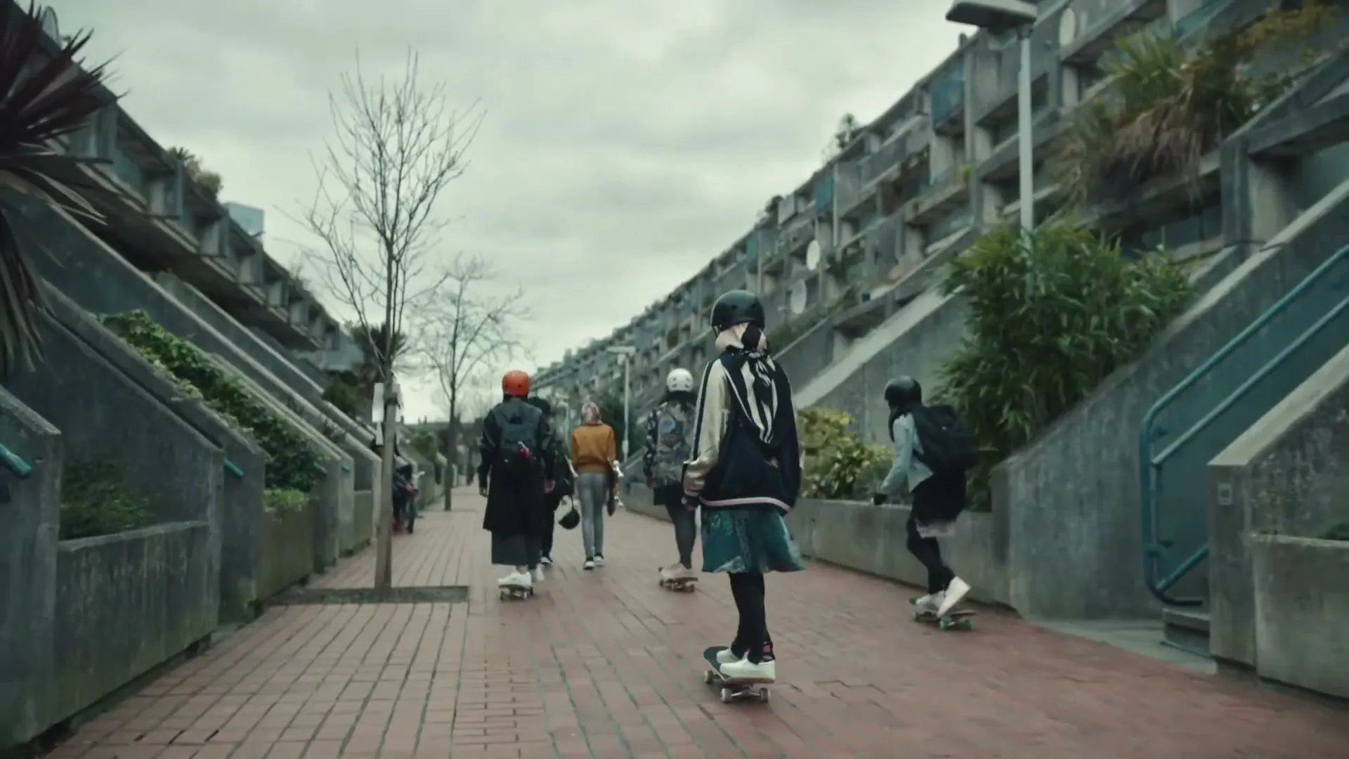 A group of diverse young people, dressed in casual streetwear and helmets, skateboard down a brick path between brutalist concrete buildings on an overcast day.