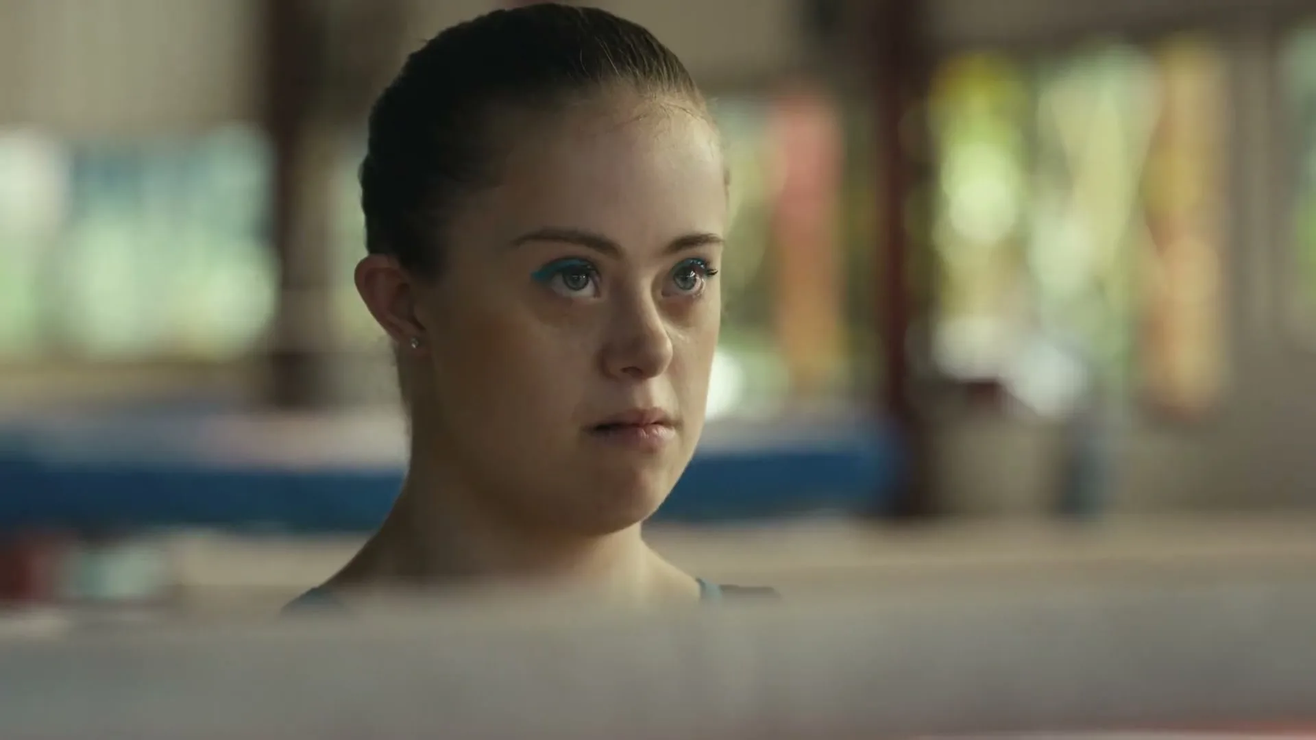A young woman with Down syndrome, wearing blue eyeliner and a blue leotard, looks intently forward in a gymnastics gym.
