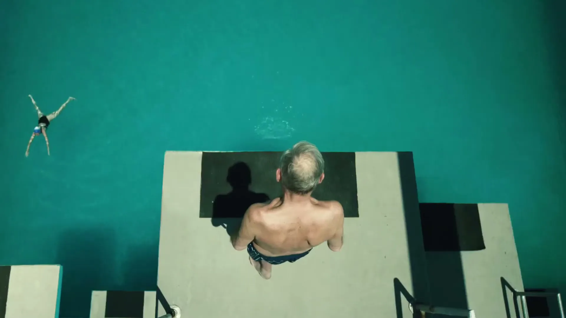 An older man in swim trunks stands on a diving board, looking down at a woman swimming in a pool, shot from directly above.