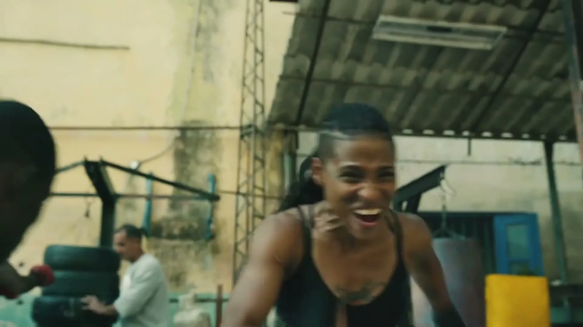 A determined female boxer with braided hair and a tank top trains intensely, gritting her teeth in an old, gritty gym.
