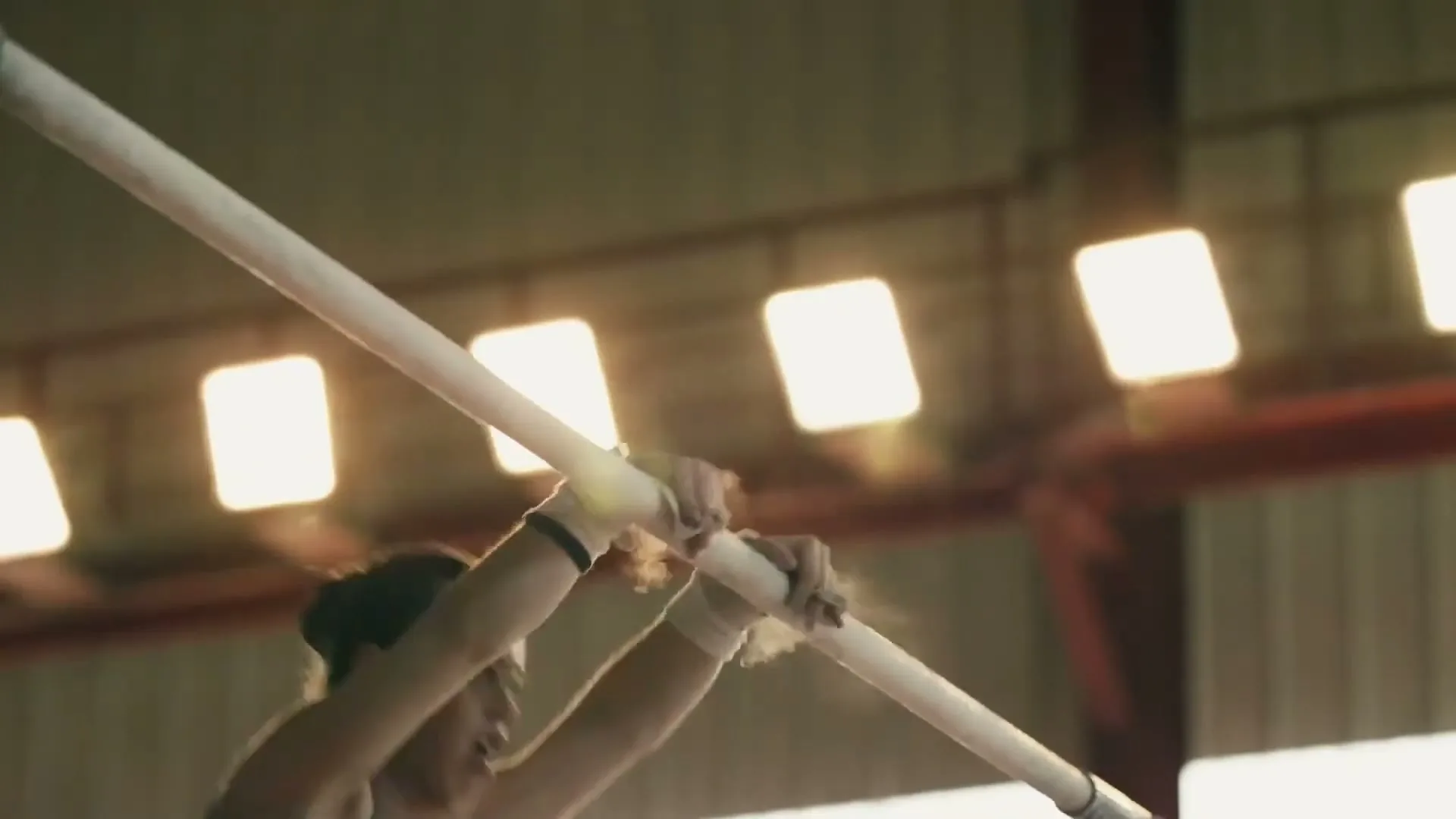A female gymnast in a blue leotard swings on uneven bars, chalk dust flying, in a brightly lit gymnasium.