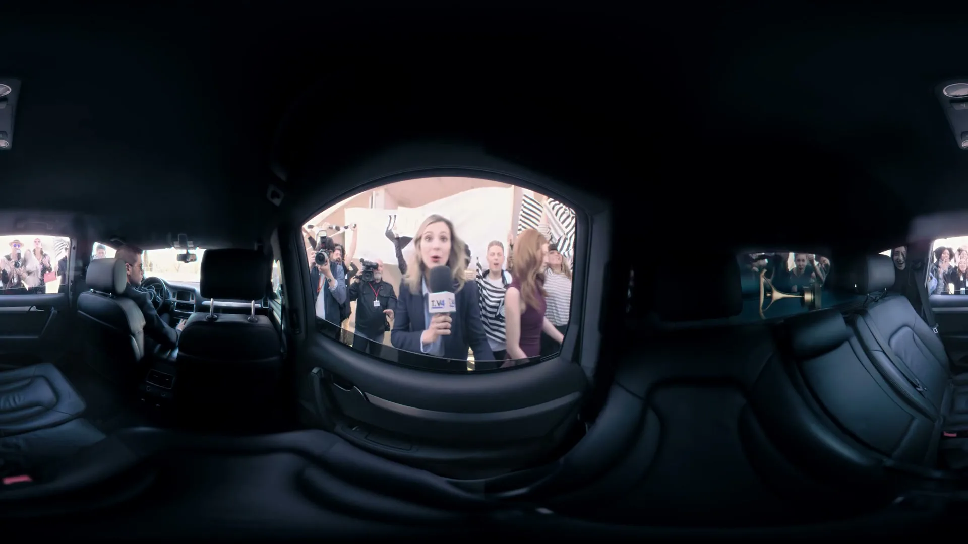 A female reporter with a microphone interviews a woman in a car, surrounded by paparazzi and fans, from inside a car.