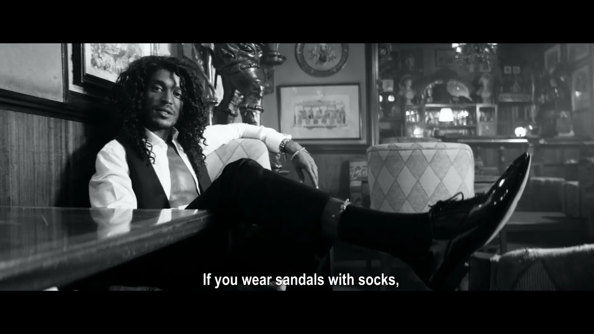 A man with long curly hair, wearing a white shirt, black vest, and black pants, sits with his feet on a table in a dimly lit, ornate room.