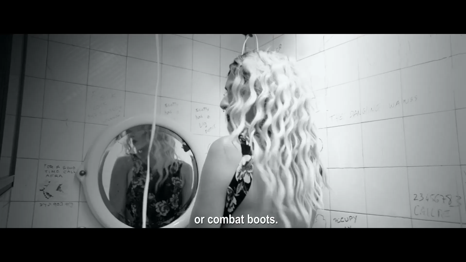 A person with long, curly blonde hair and a floral top looks over their shoulder at their reflection in a round mirror in a tiled bathroom.