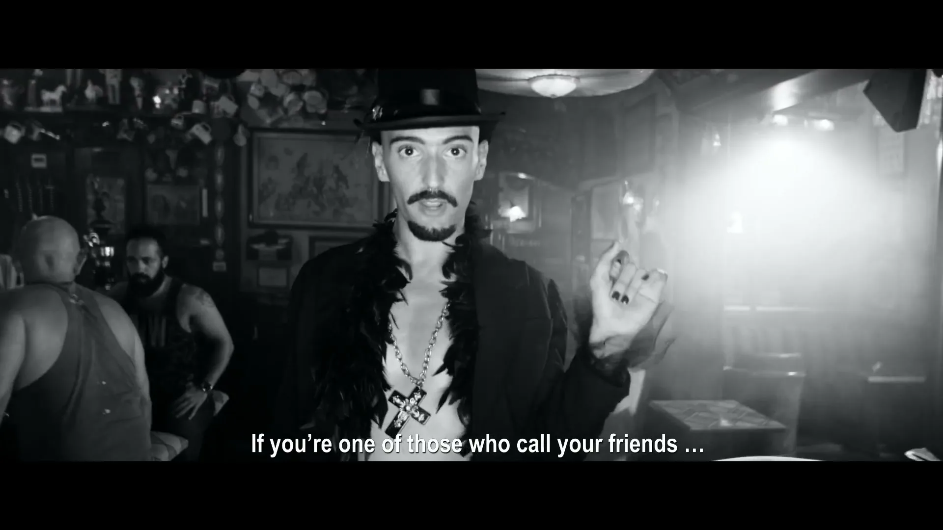 A man in a top hat and feather boa, with a cross necklace, speaks directly to the camera in a dimly lit bar, gesturing with his hand.