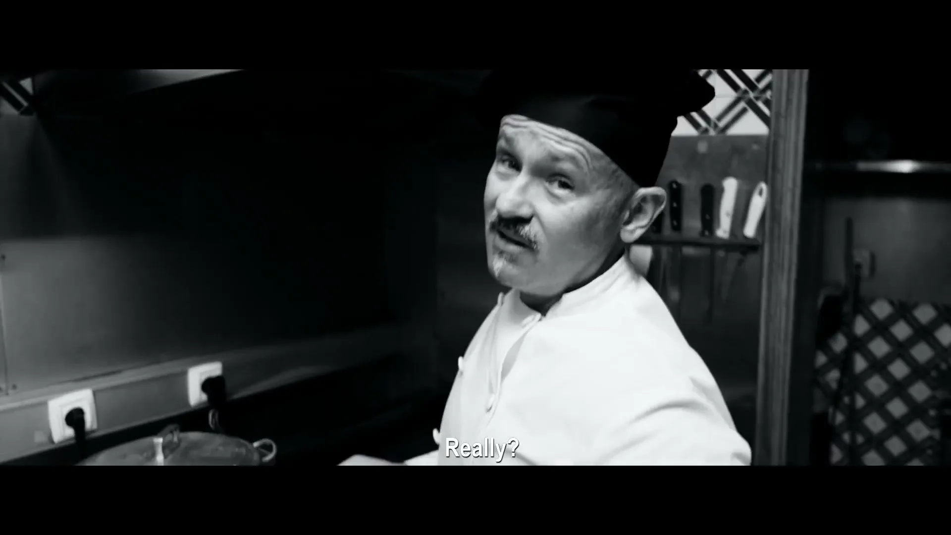 A male chef in his 50s, wearing a chef's hat and jacket, looks directly at the camera with a questioning expression in a dimly lit kitchen.