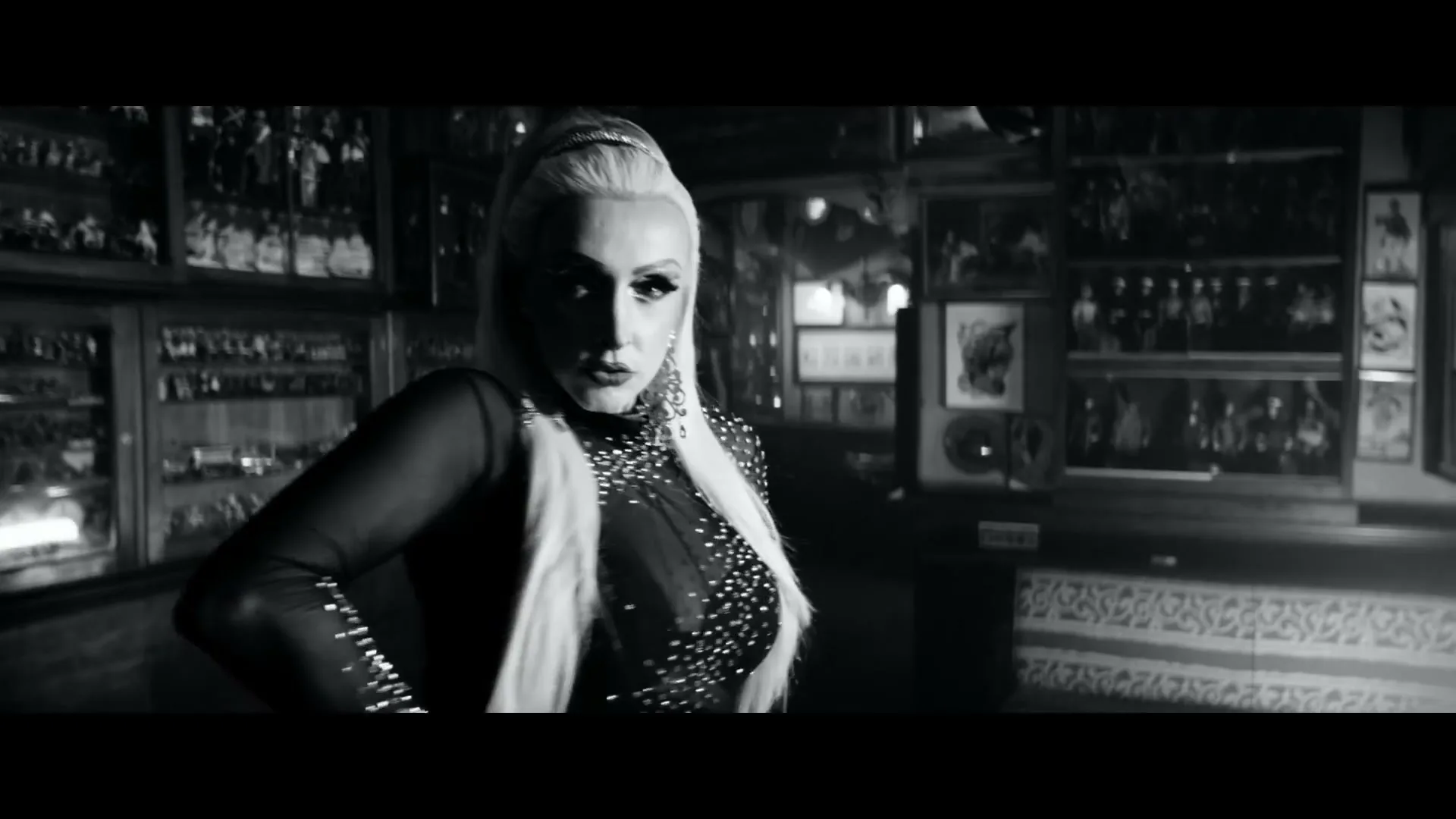 A drag queen with blonde hair and a sparkly outfit poses confidently in a dimly lit room filled with display cases and framed pictures.