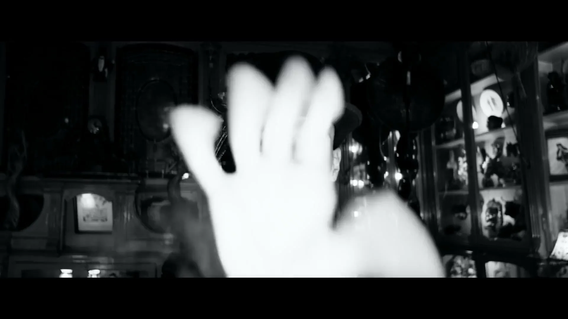 A man in a top hat and dark clothes gestures with his hand, speaking to the camera in a dimly lit, cluttered room.