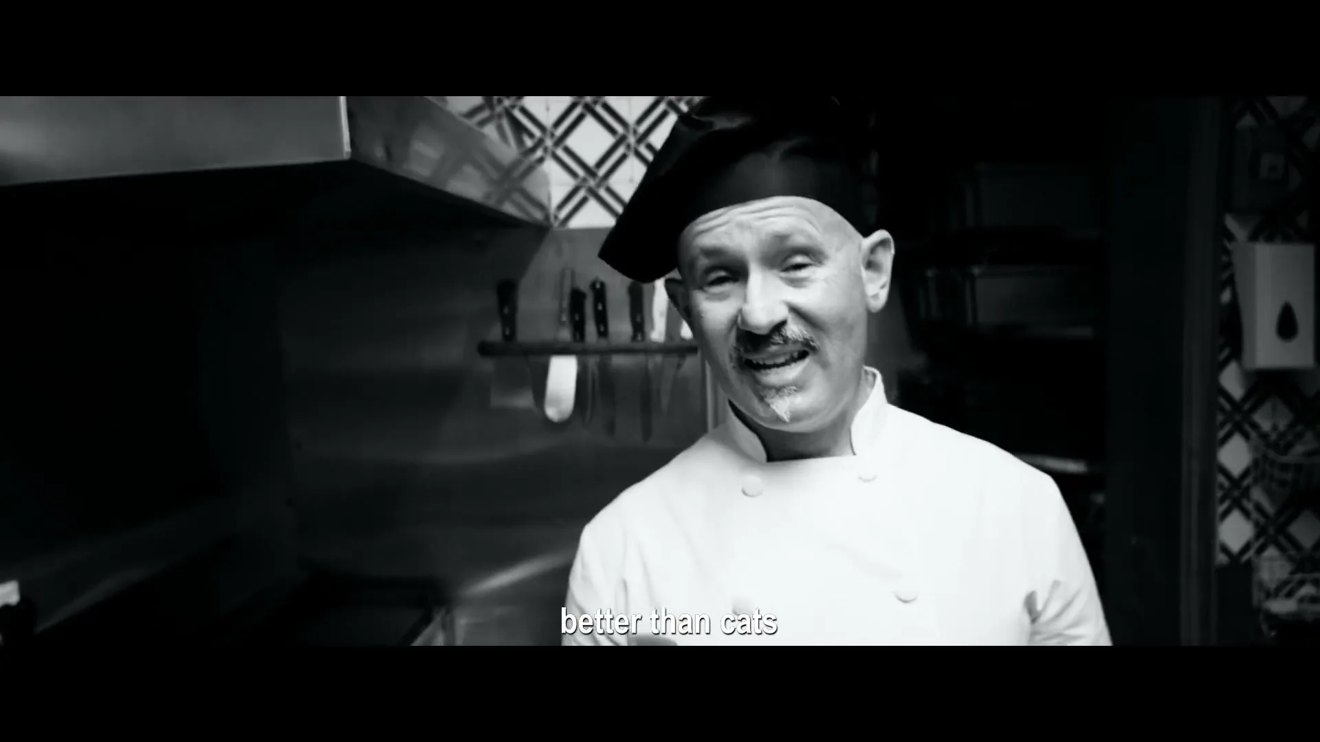 A male chef in his 50s, wearing a white chef's coat and black beret, smiles at the camera in a black and white shot inside a restaurant kitchen.