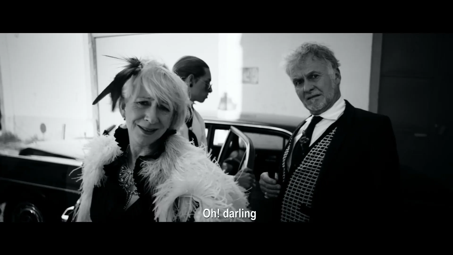 A woman in a feathered boa and a man in a suit stand next to a car, looking at the camera, in a black and white shot.