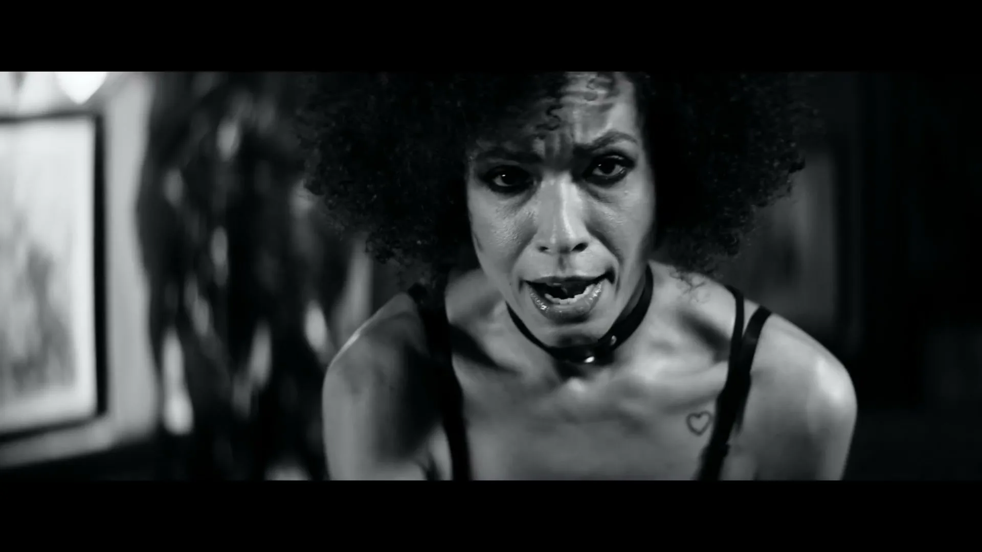 A woman with dark curly hair and a choker looks intensely at the camera in a black and white shot.
