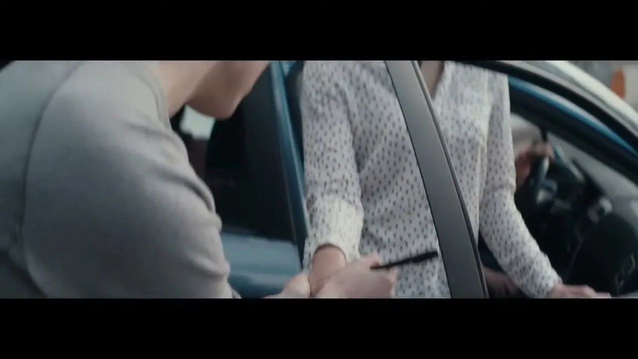 A young man in a grey hoodie hands a black pen to a young woman in a white polka dot shirt, who is sitting in a car.
