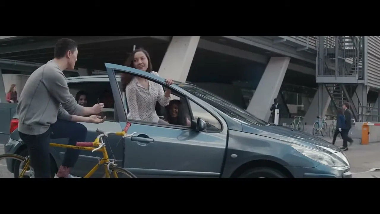 A young man on a bicycle hands a drink to a young woman in a car, while other people are visible in the car and background, in an urban setting.