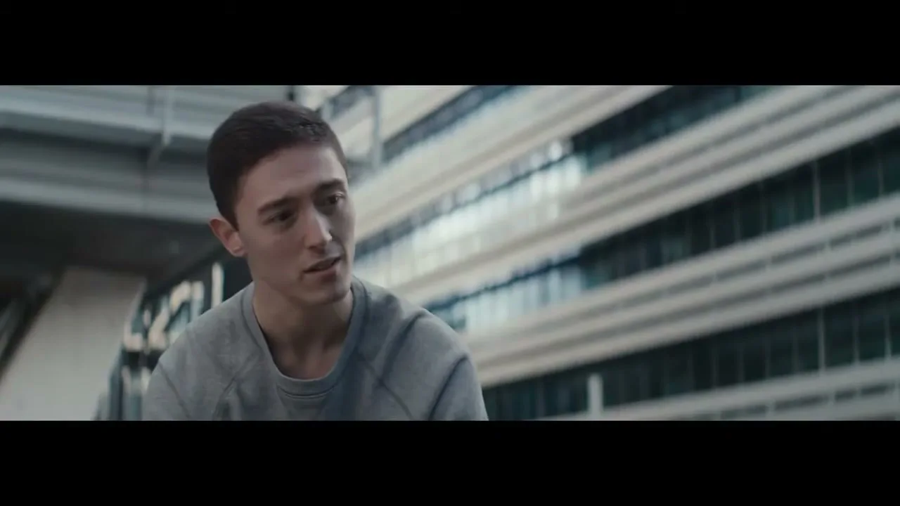 A young man with short dark hair and a grey sweatshirt looks to the right, with a modern building in the background.