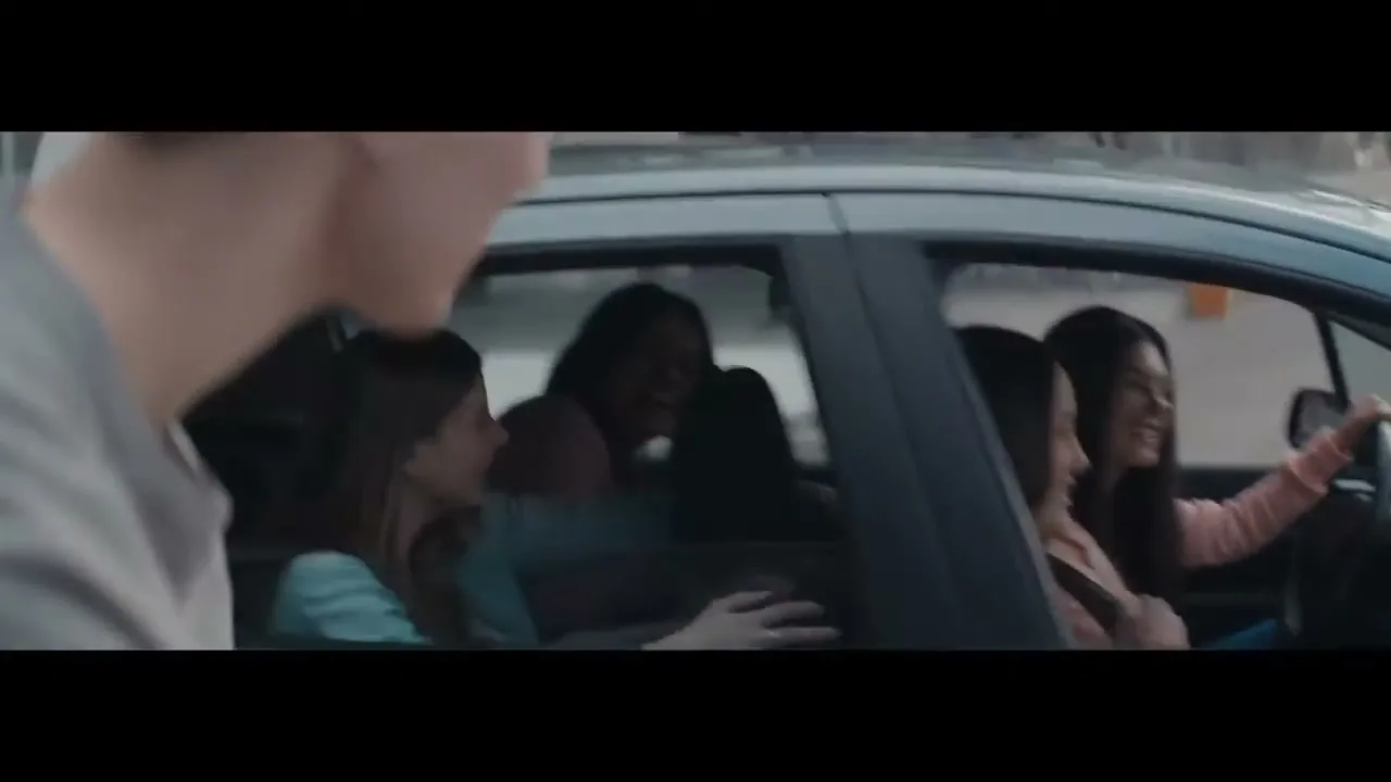 A young man in a grey t-shirt talks to a group of young women in a car, who are laughing and smiling, on a city street during the day.