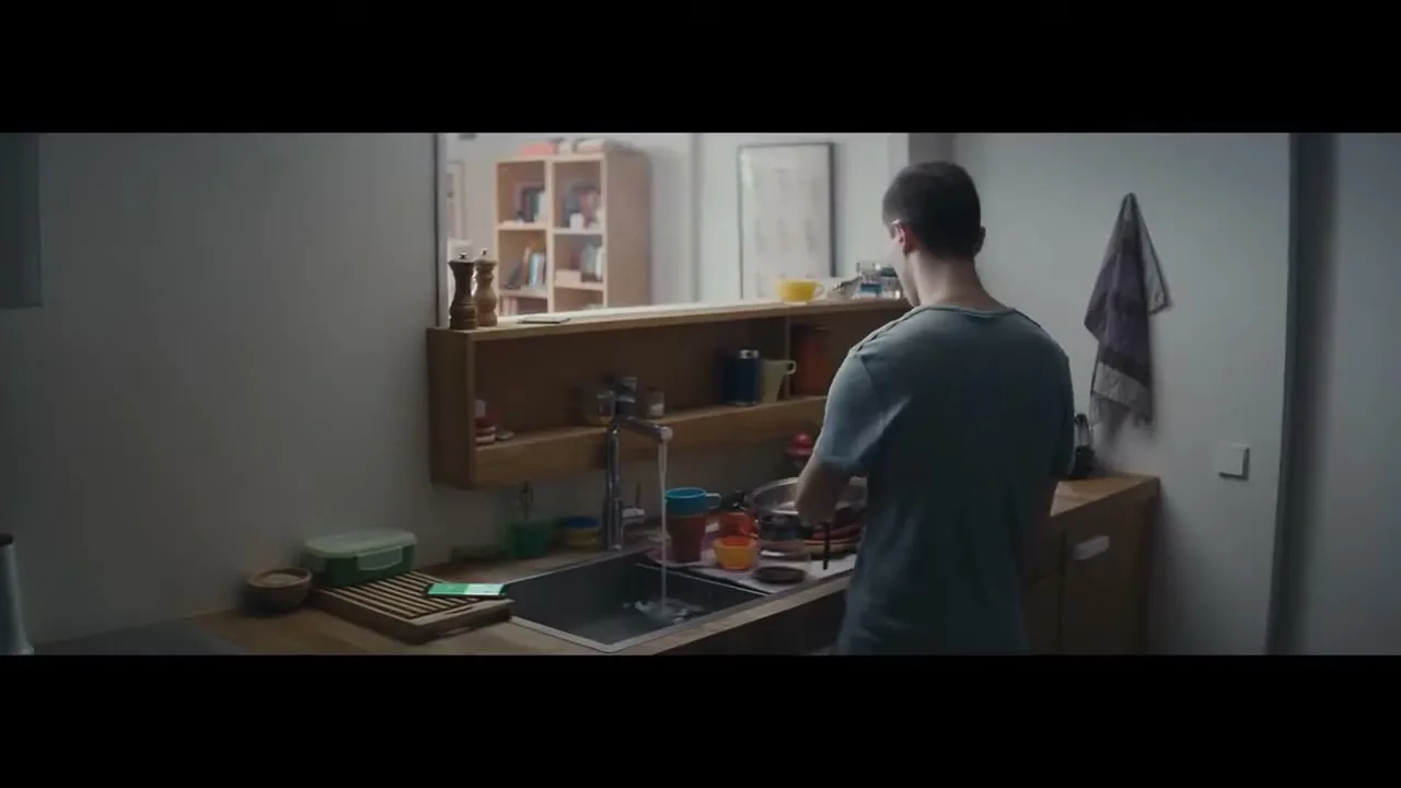 A young man in a grey t-shirt washes dishes in a kitchen sink, with a mirror reflecting shelves behind him.
