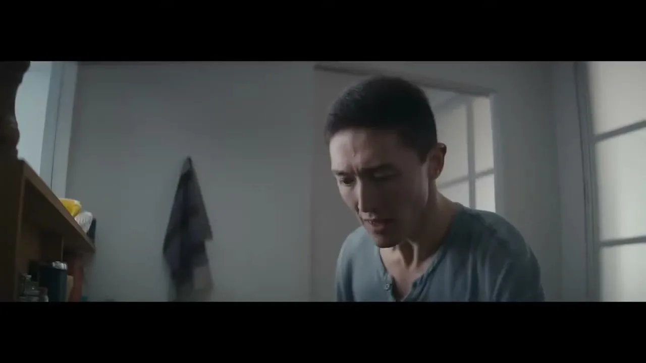 A young man with short dark hair and a blue shirt looks down with a pained expression in a dimly lit room.